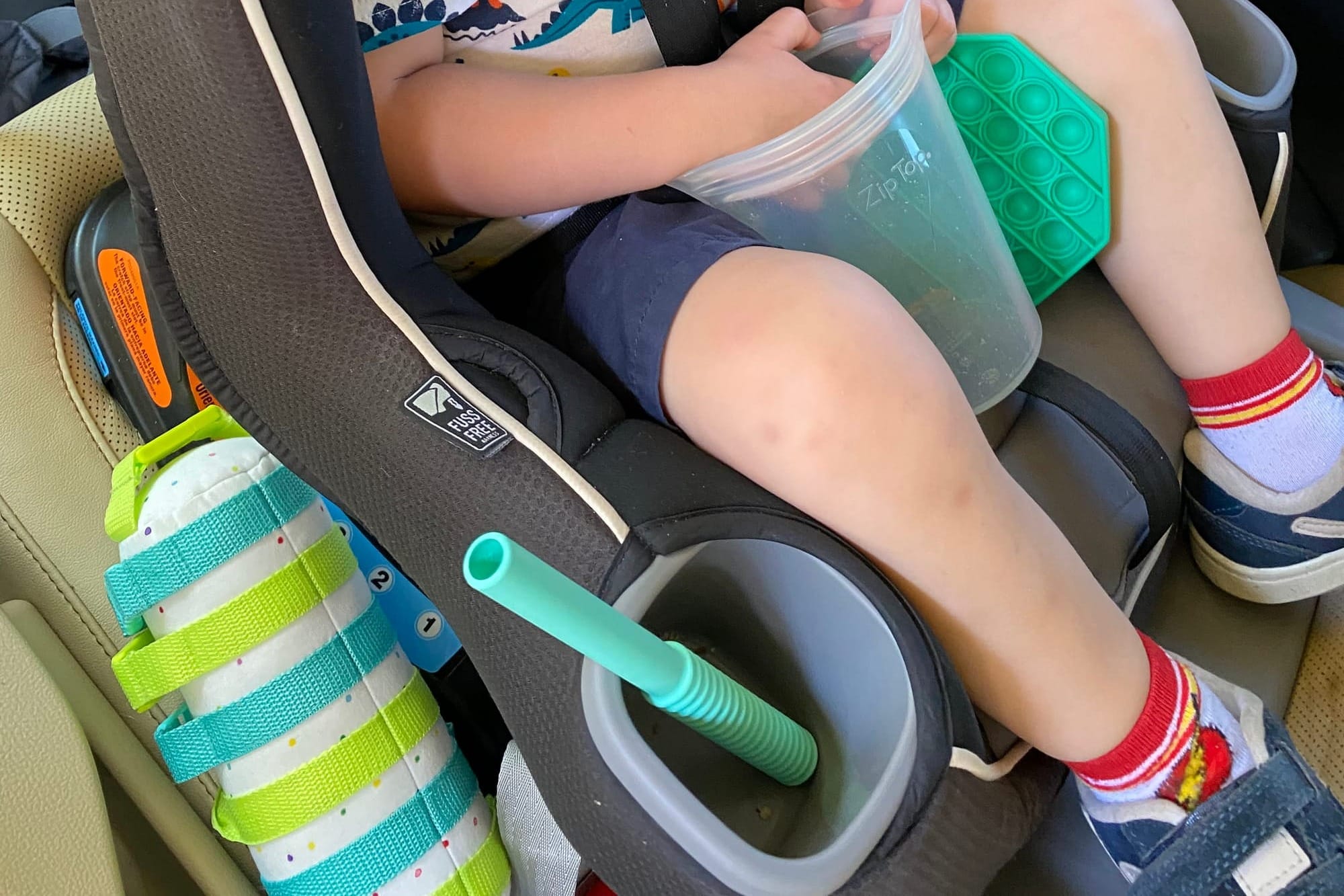 Toddler in a car seat holding a snack bag during a road trip, with a pop-it toy and teether