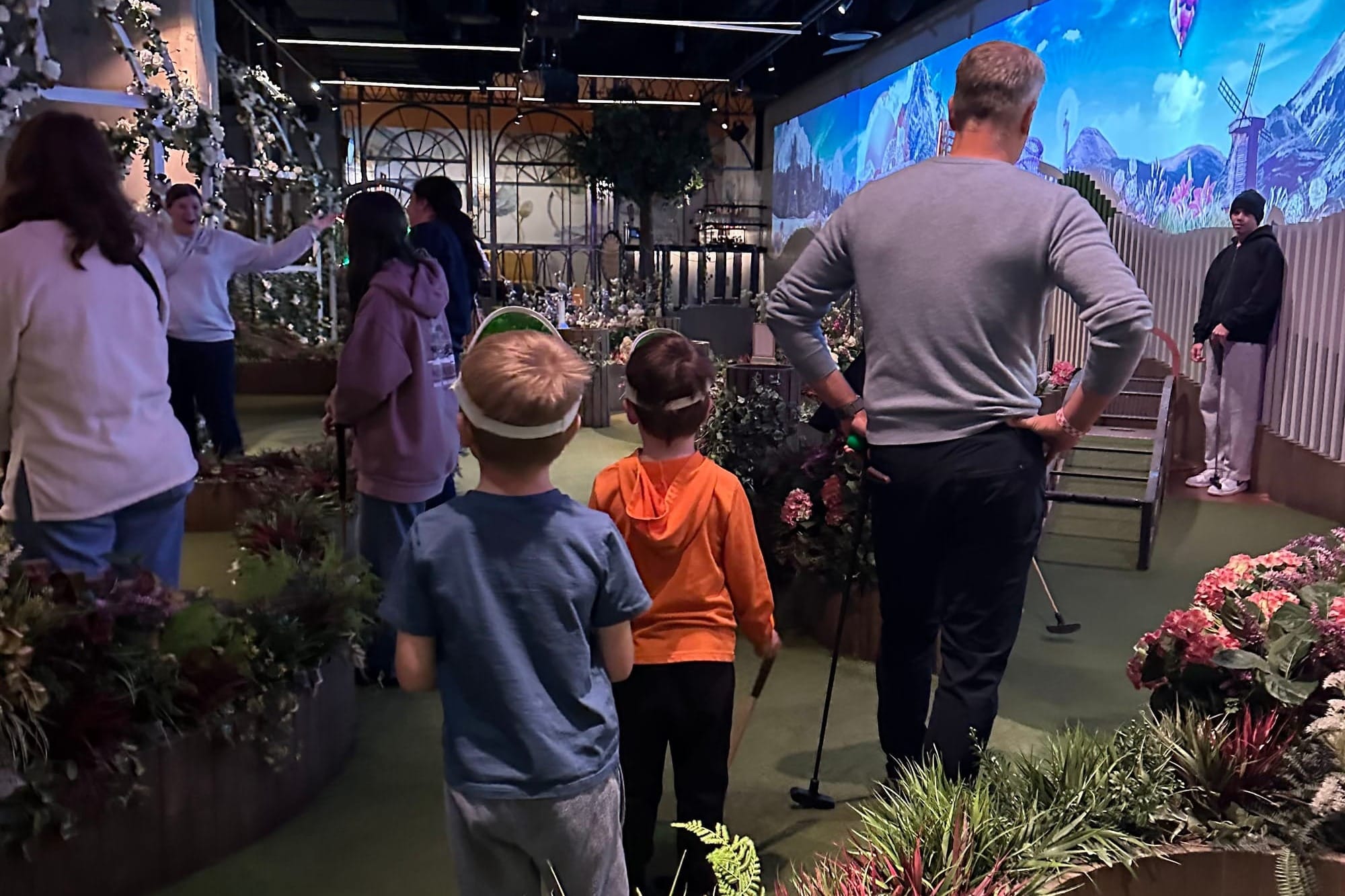Grandfather and two young kids in matching visors lining up a shot at an indoor mini golf course decorated with flowers and a mural backdrop