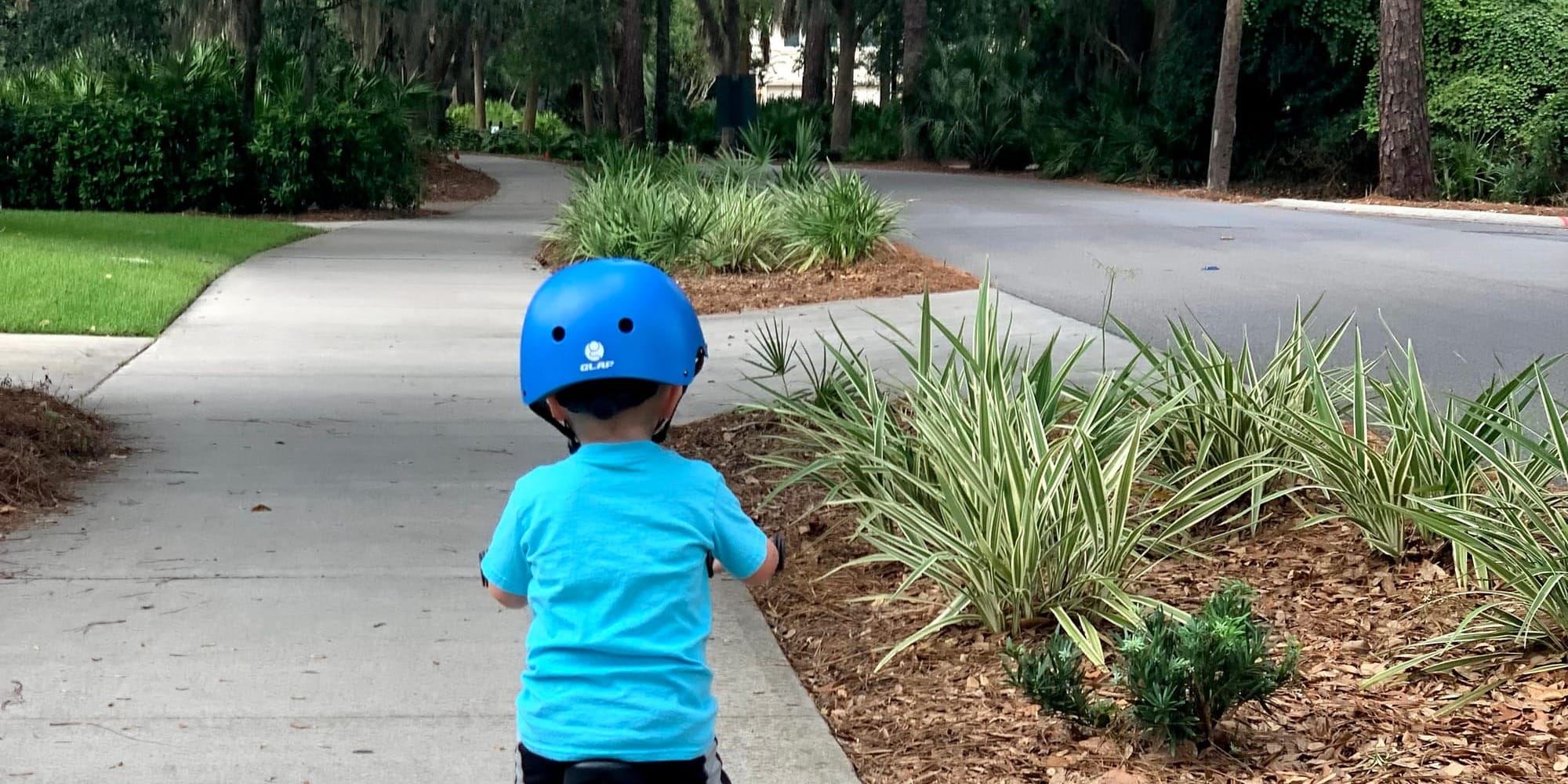 A child in a blue helmet rides a bike along a shady paved path in Hilton Head, SC lined with greenery.