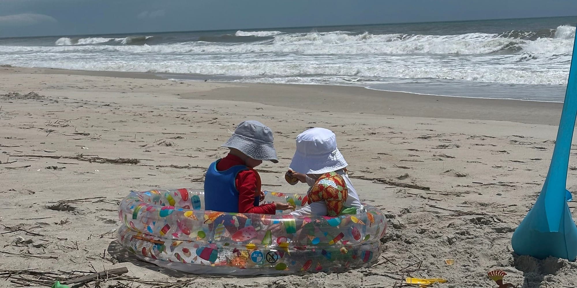 Two young children in sun hats sit in an inflatable pool on the sand with waves breaking behind them in Surf City, NC.