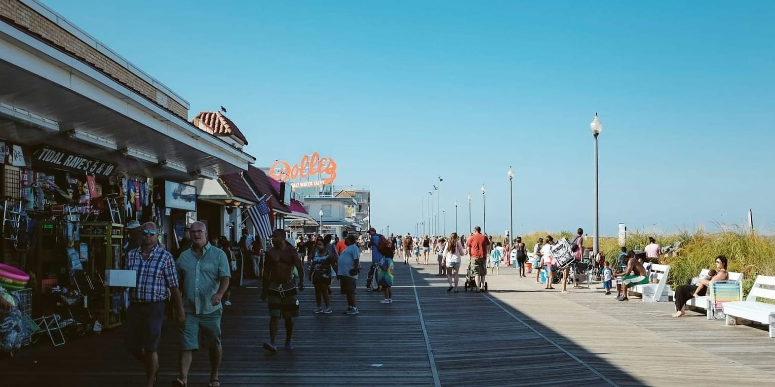 Families with stroll along a wooden boardwalk in Rehoboth Beach lined with shops on a sunny day.