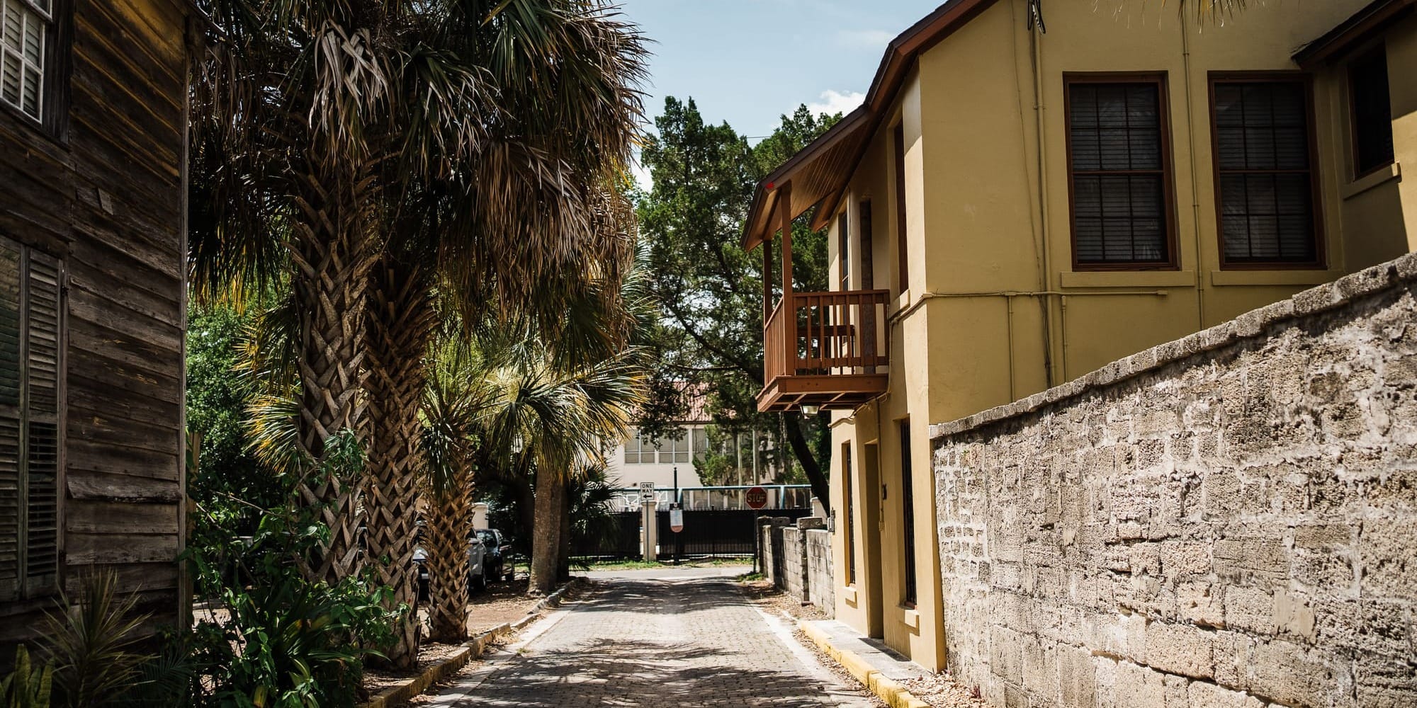 A narrow historic St. Augustine street lined with old buildings, a stone wall, and palm trees.