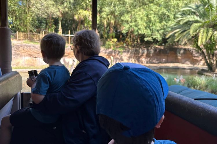 Grandmother and grandson sharing binoculars on a safari ride, scanning the riverbank for animals with two more kids in the row ahead