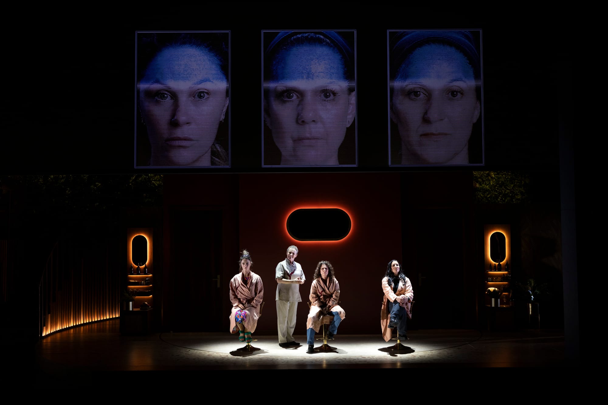 Four women sit facing the audience beneath three large projected female faces in Losing Face.