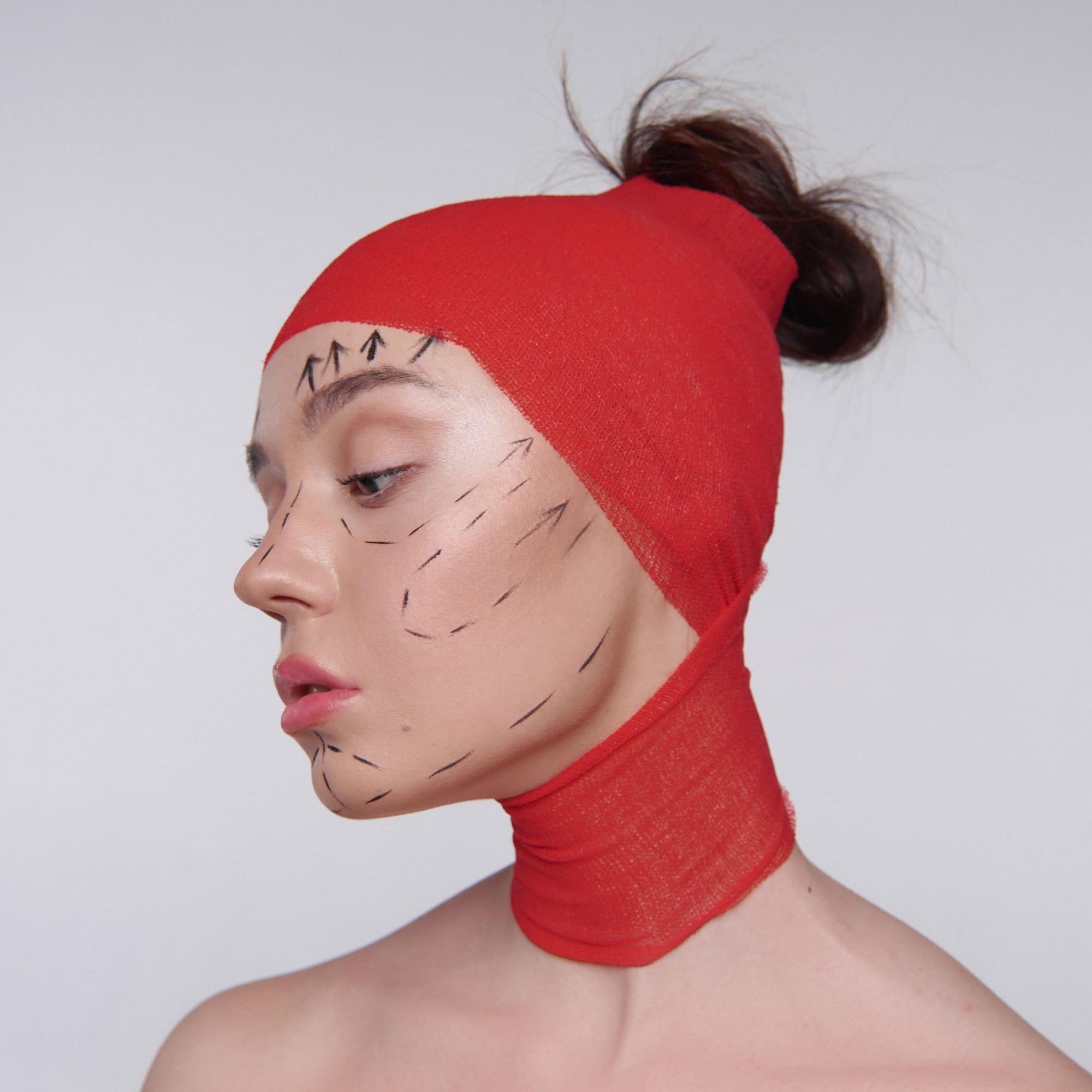 Young woman in profile wearing a red surgical head wrap, with cosmetic surgery markings drawn across her face.