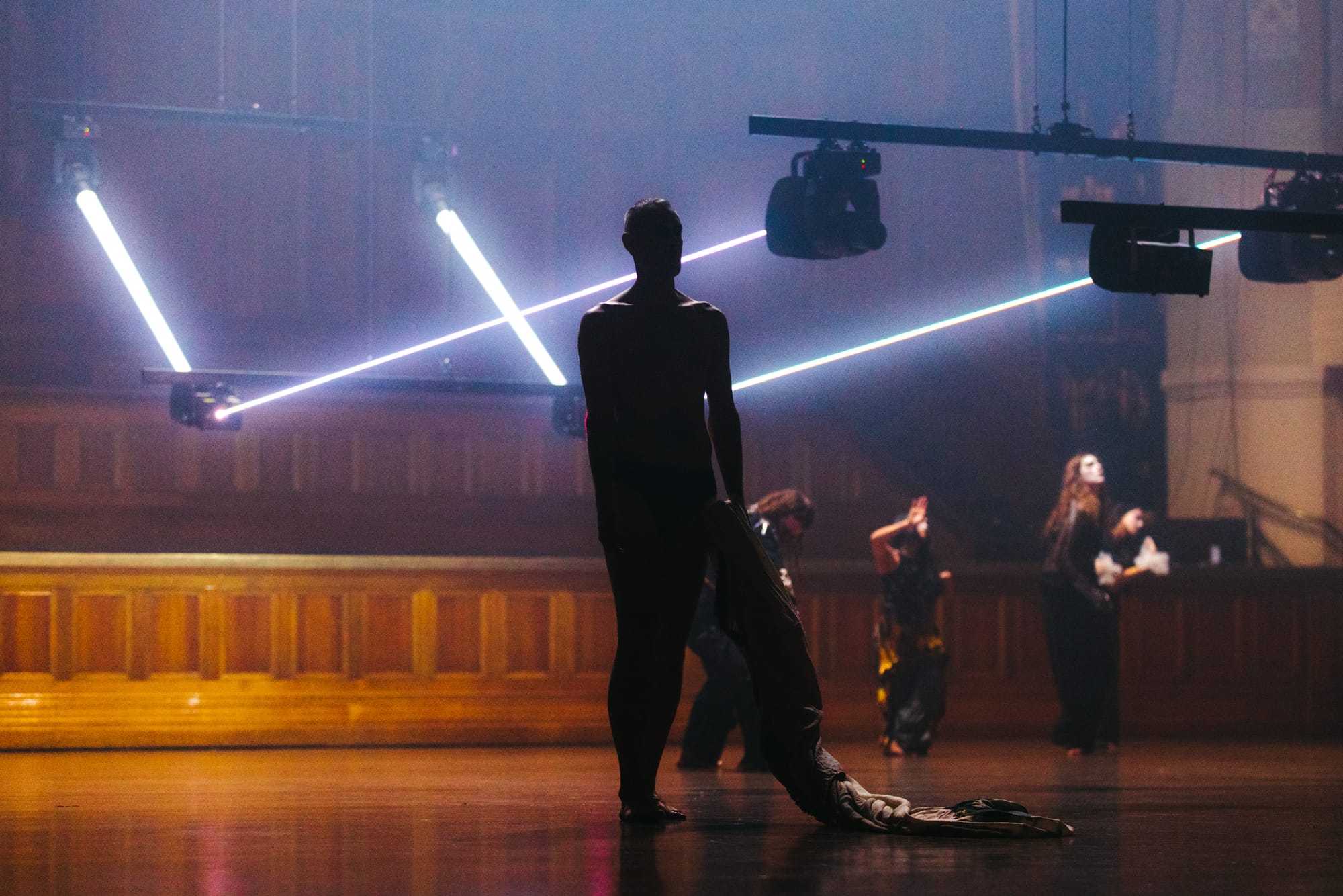 A silhouetted dancer stands on the timber floor of Melbourne Town Hall holding a trailing costume piece, while other performers move in the distance beneath bright crossing laser beams in Chunky Move’s Time Remaining.
