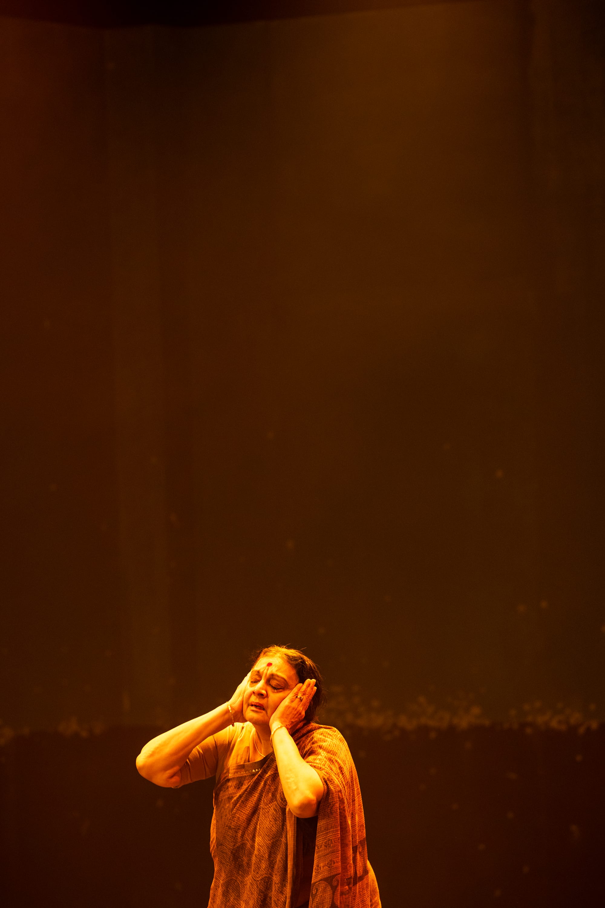 Gowrie stands under warm amber light with her hands raised to her face.