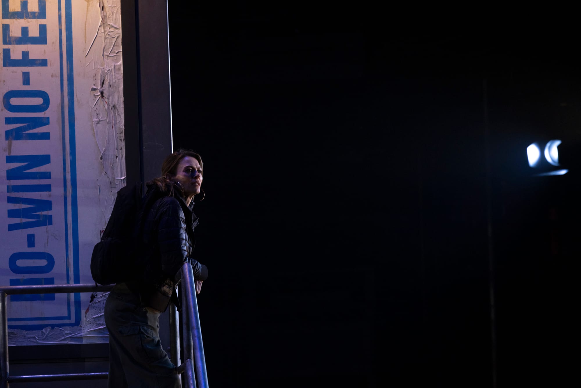 Pamela leans forward at the edge of the staircase, partially lit against the darkness of the warehouse setting.
