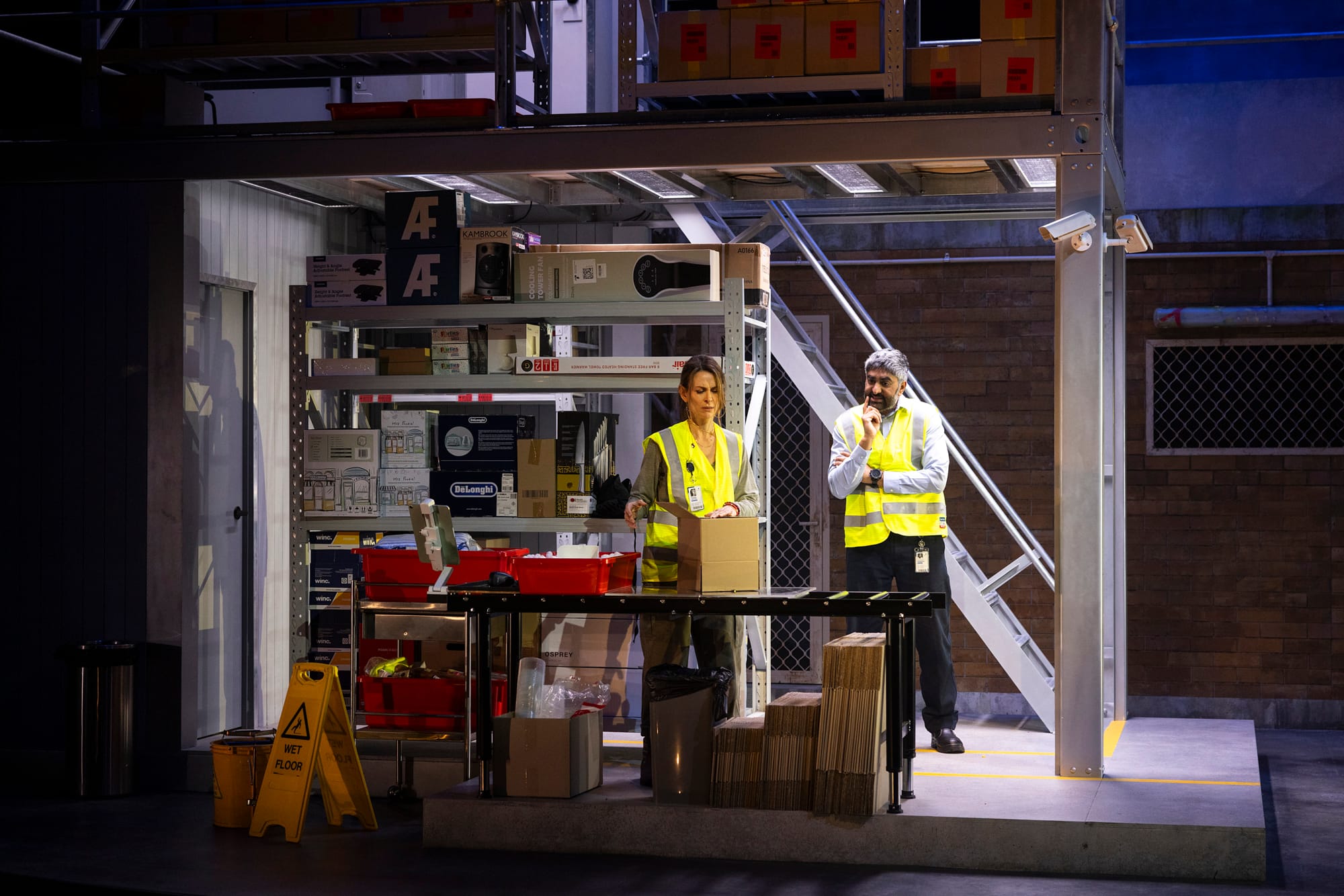 Pamela and Sanjeeva, wearing high-visibility workwear, stand together inside the warehouse beneath the metal staircase of the two-storey set.