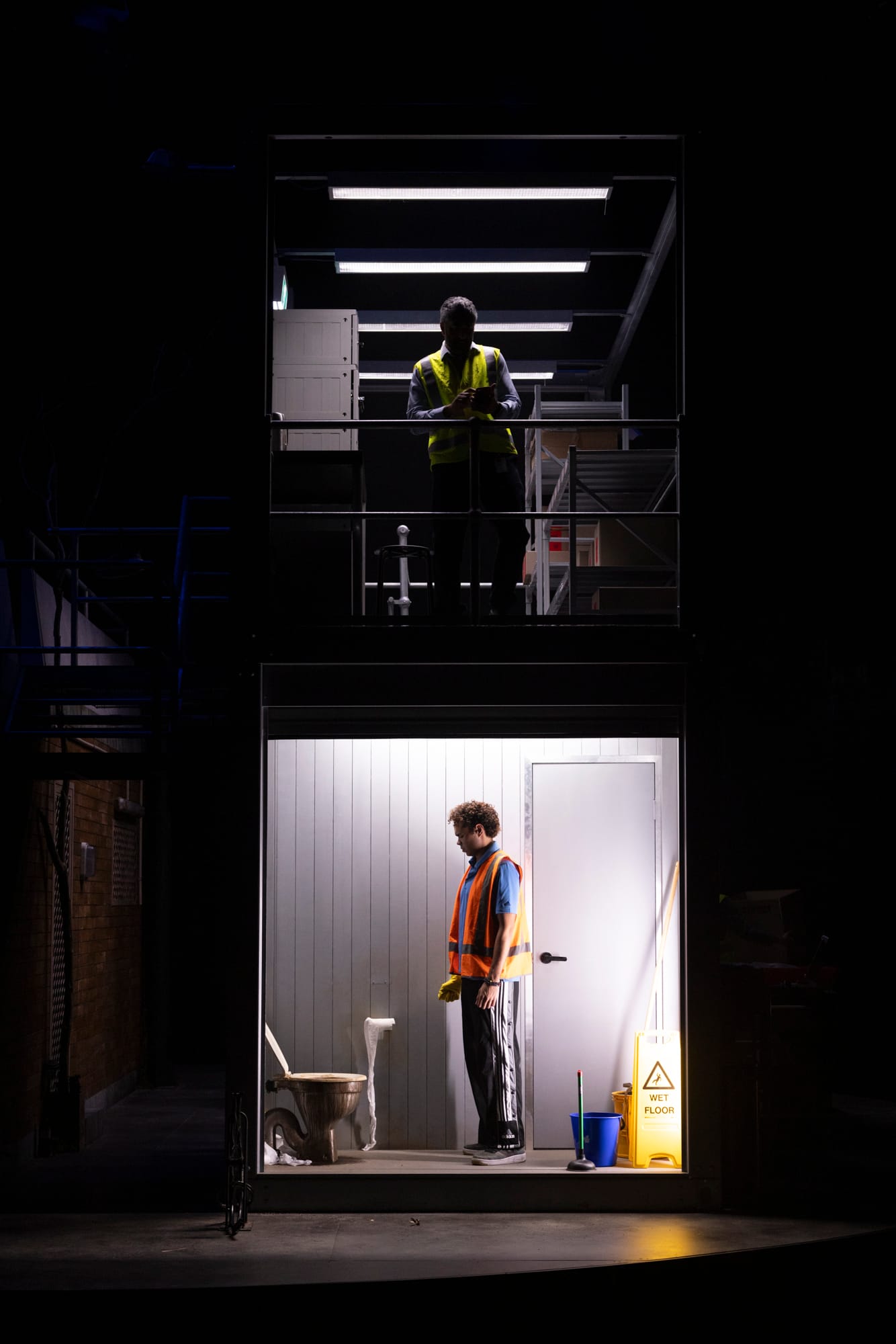Jedediah stands beside a toilet in the brightly lit lower-level utility room, while his supervisor Sanjeeva stands above him on the warehouse level in high-visibility clothing.