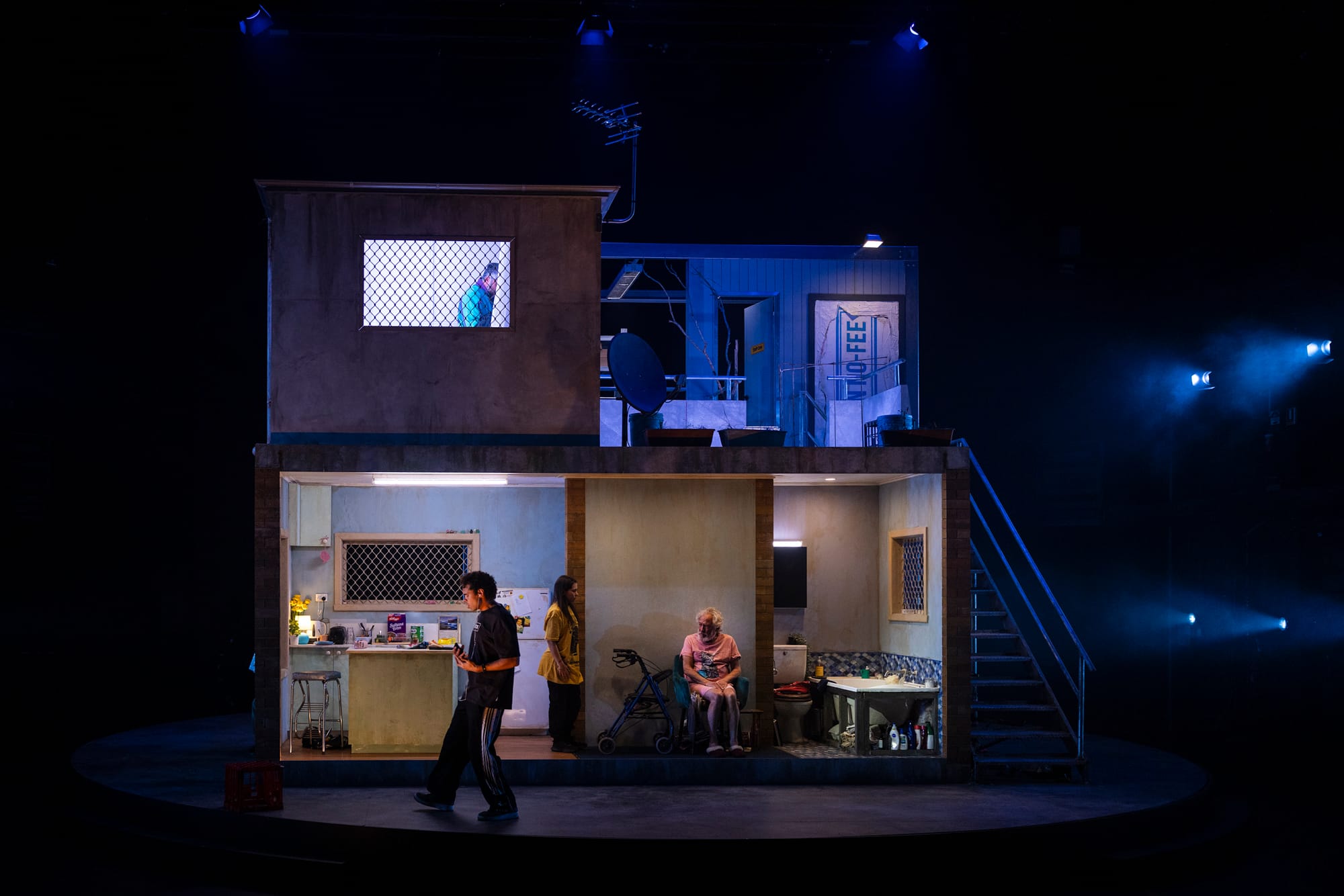 Jedediah stands outside the brightly lit house set while Rae stands inside the kitchen and Pop sits nearby in his chair, all framed within the larger dark stage.