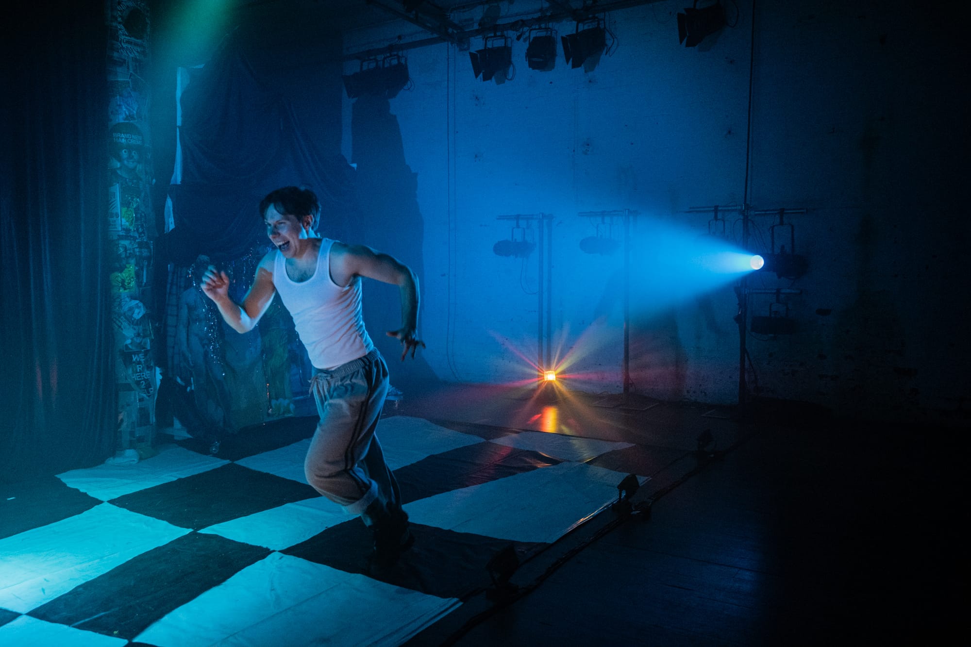 Ludomyr Kemp-Mykyta as Tyler runs across a black-and-white checkerboard stage, caught mid-stride under blue theatrical lighting.