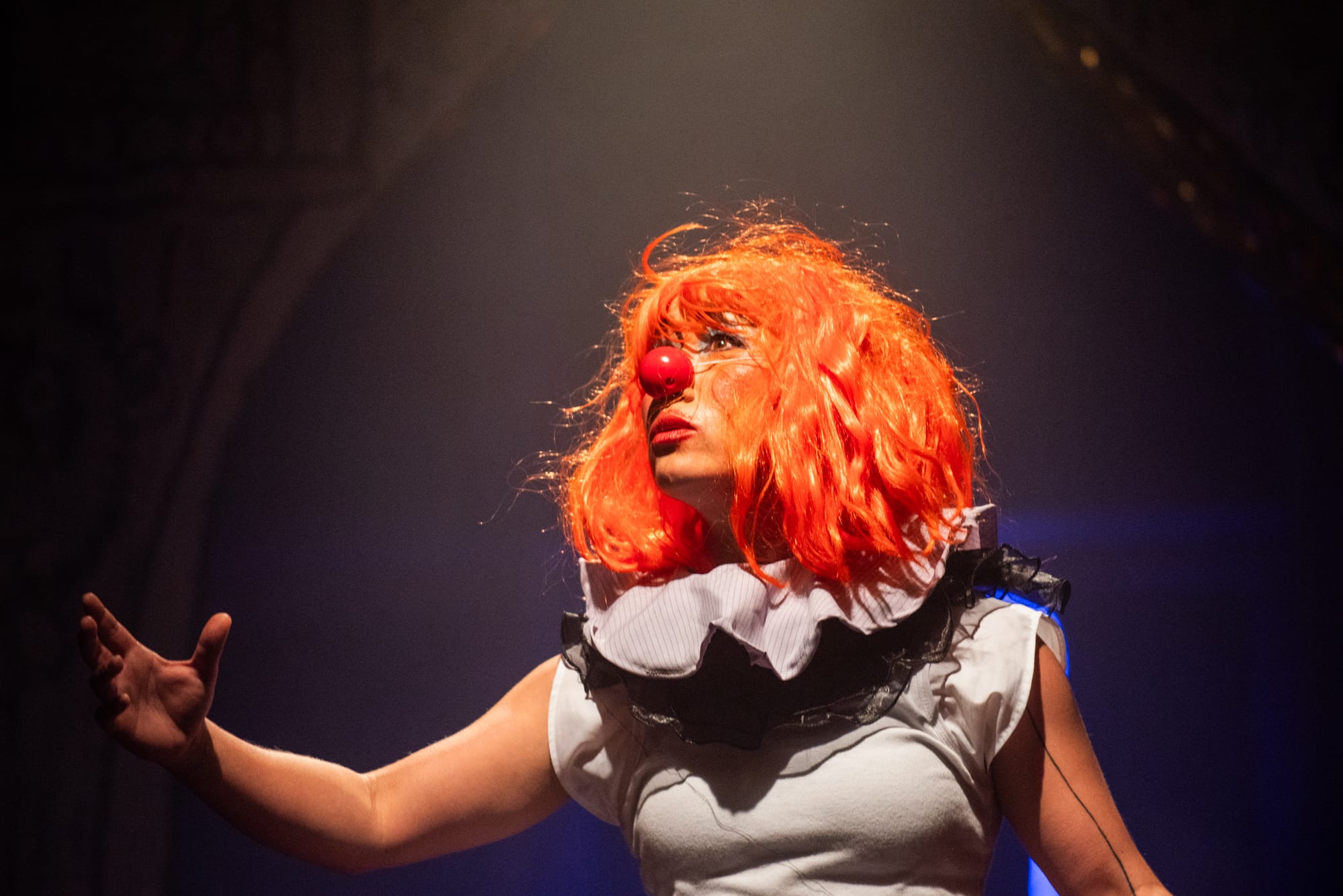 AYA performs in a vivid orange wig and exaggerated clown makeup, gesturing with one hand against the dark theatrical set.