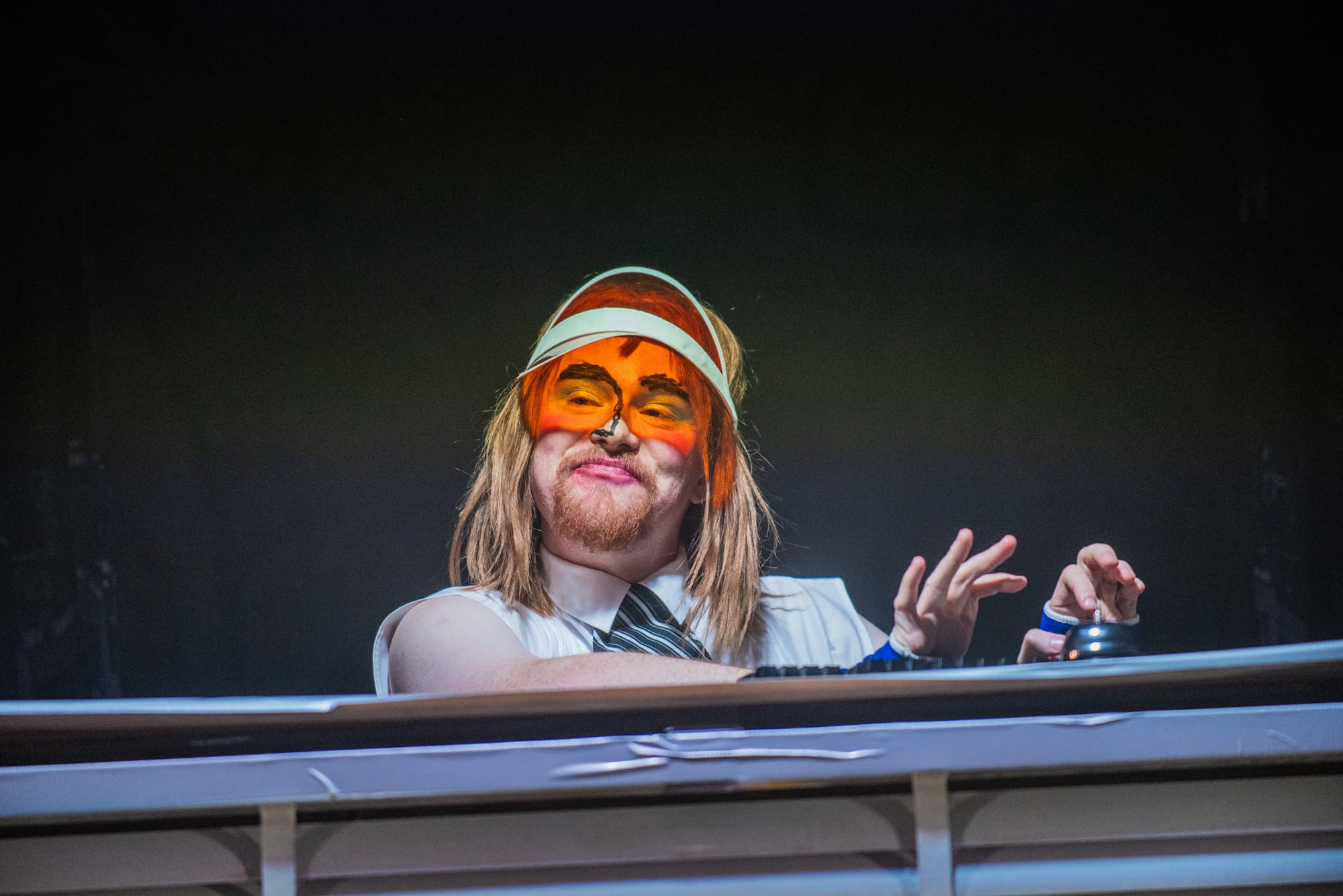 Harry McGee as a welfare clerk peers down from behind horizontal blinds, wearing an orange visor as performers queue below in the production’s absurdist Centrelink-like office.