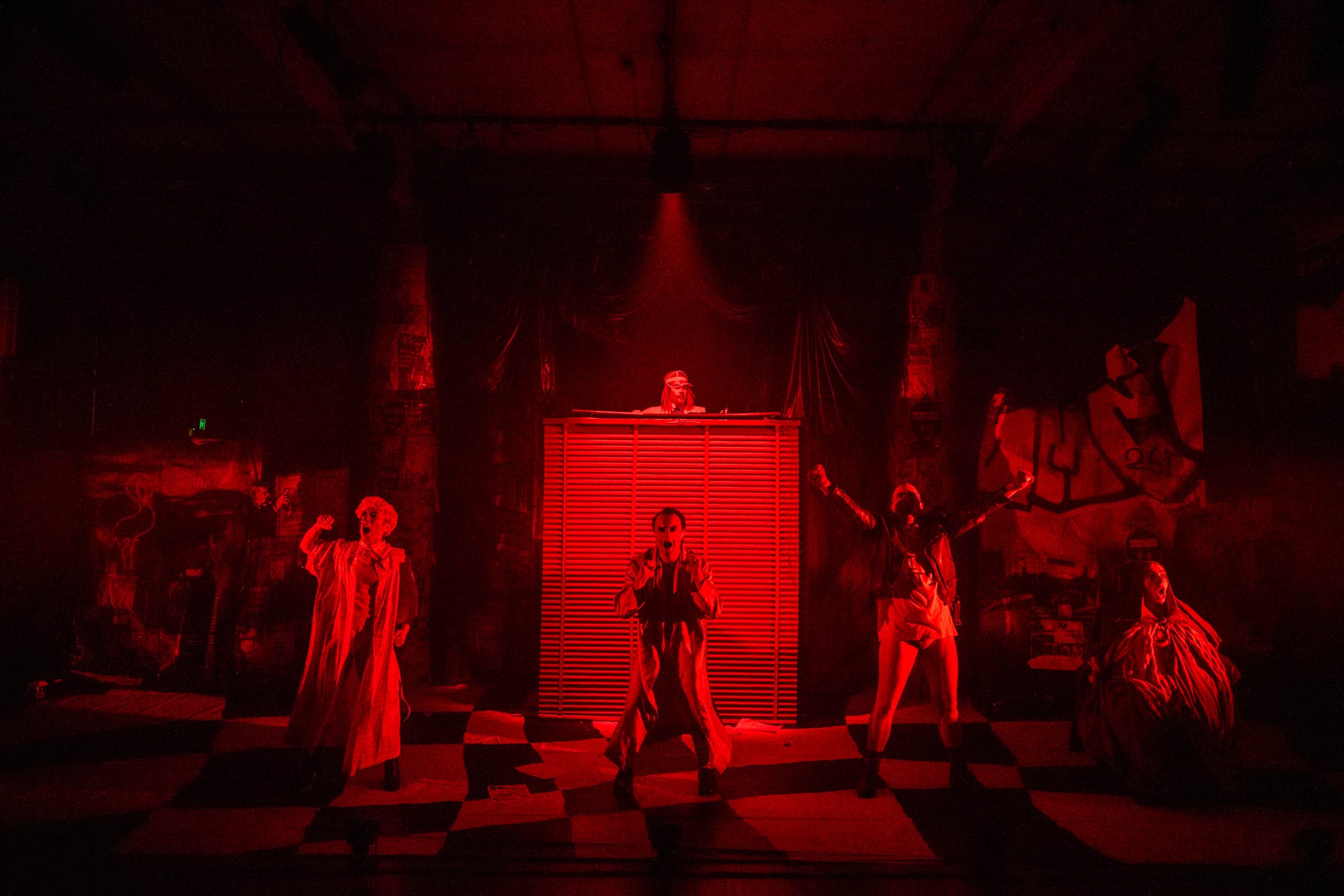 The Marginalia ensemble spread across the stage beneath intense red lighting, their bodies caught in exaggerated poses amid the production’s ramshackle set.