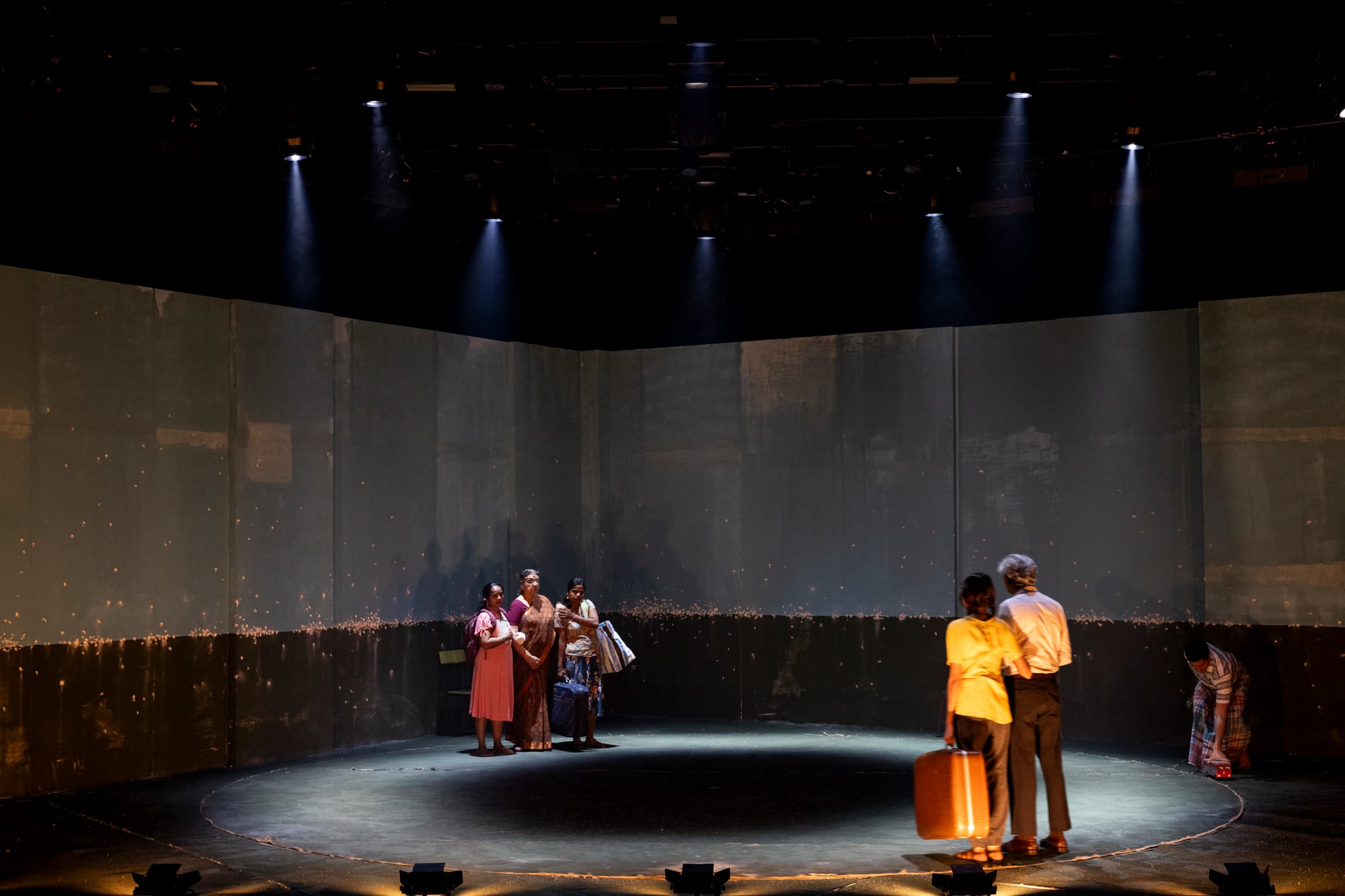 Three women embrace at one side of the revolve as another group gathers across the stage beneath a darkened blue set.
