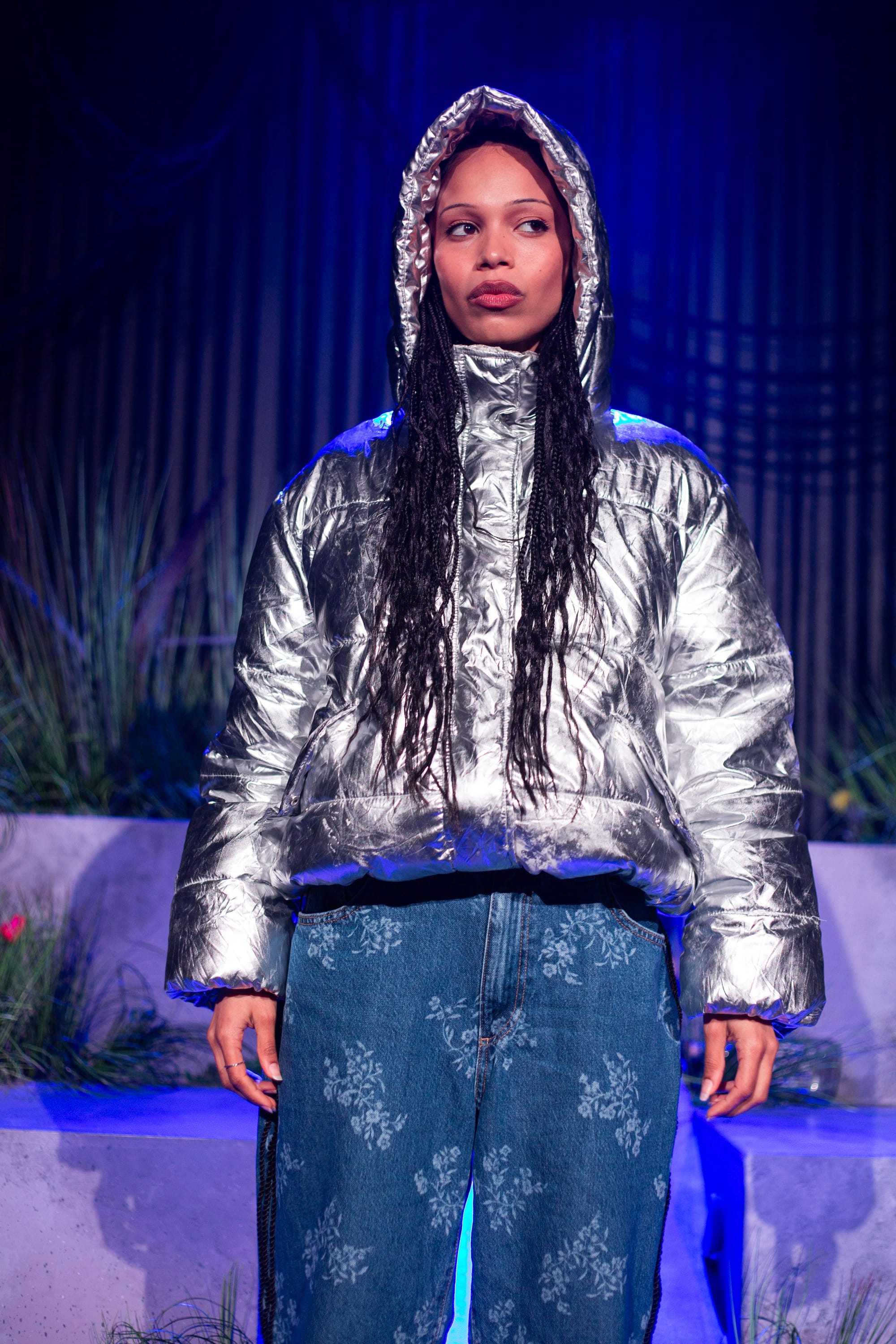 Lucy Ansell stands centre stage wearing a silver jacket against a blue-lit backdrop in Funeral Flowers.