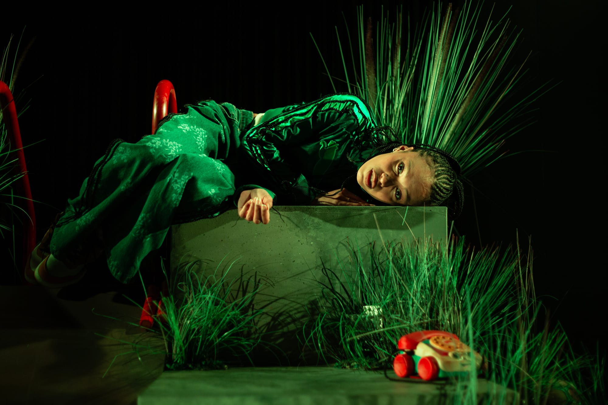 Lucy Ansell lies curled on a concrete platform surrounded by flowers and foliage under green stage lighting in Funeral Flowers.