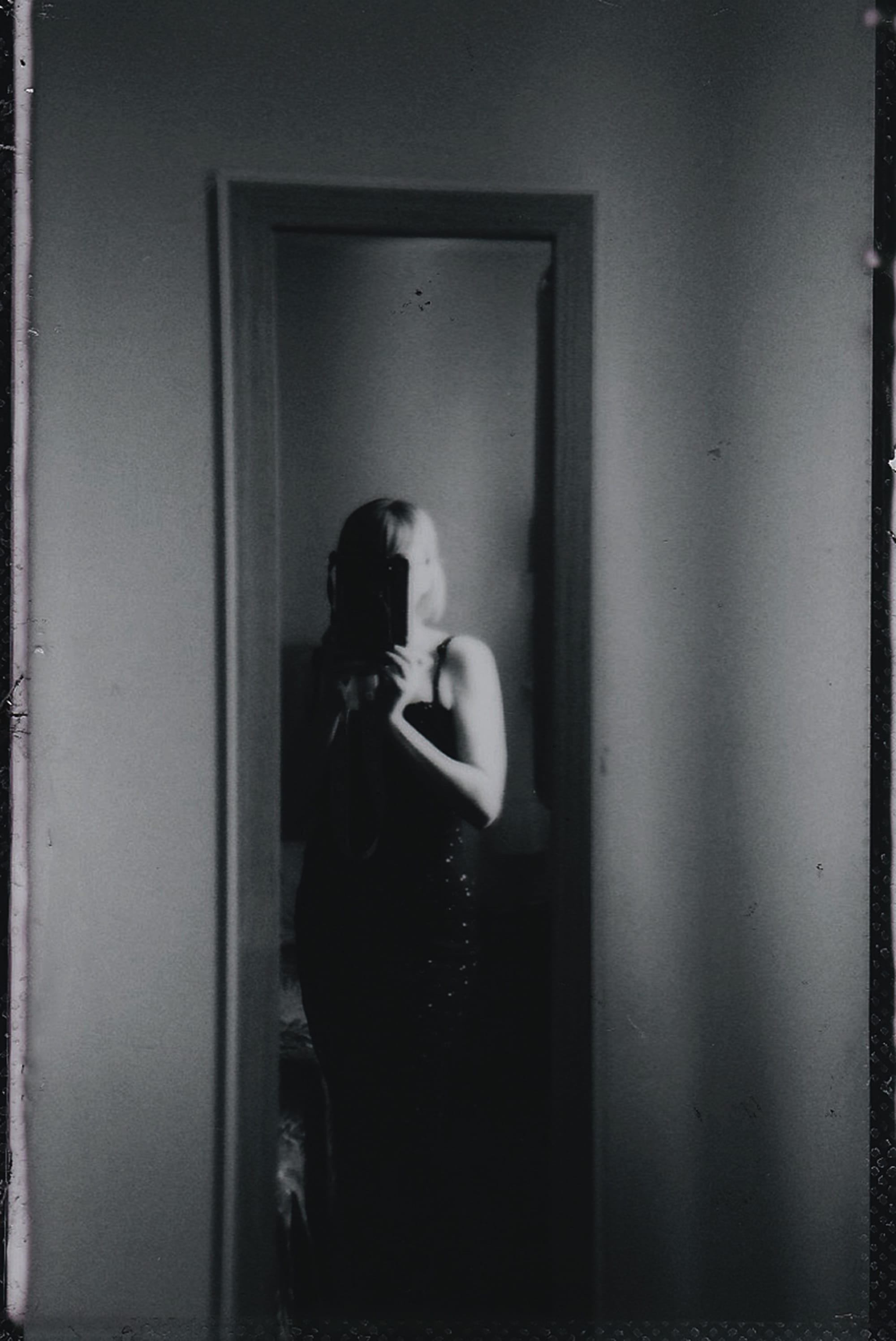 A woman standing partly in shadow in a doorway, framed by the dark interior around her.