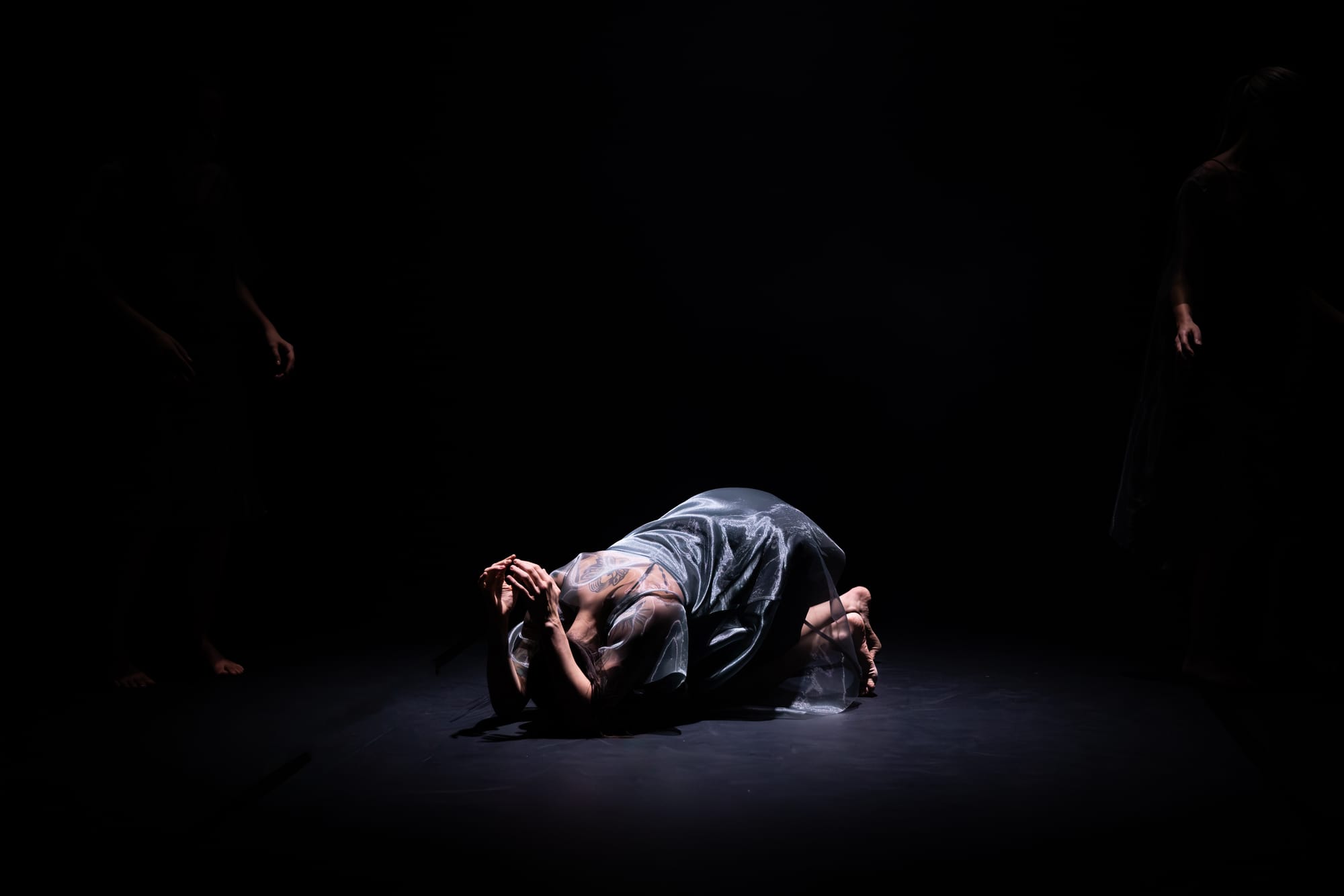 Rachel Coulson curls low to the floor, isolated in a pool of light against the darkened stage in 7 Dances for 7 People.