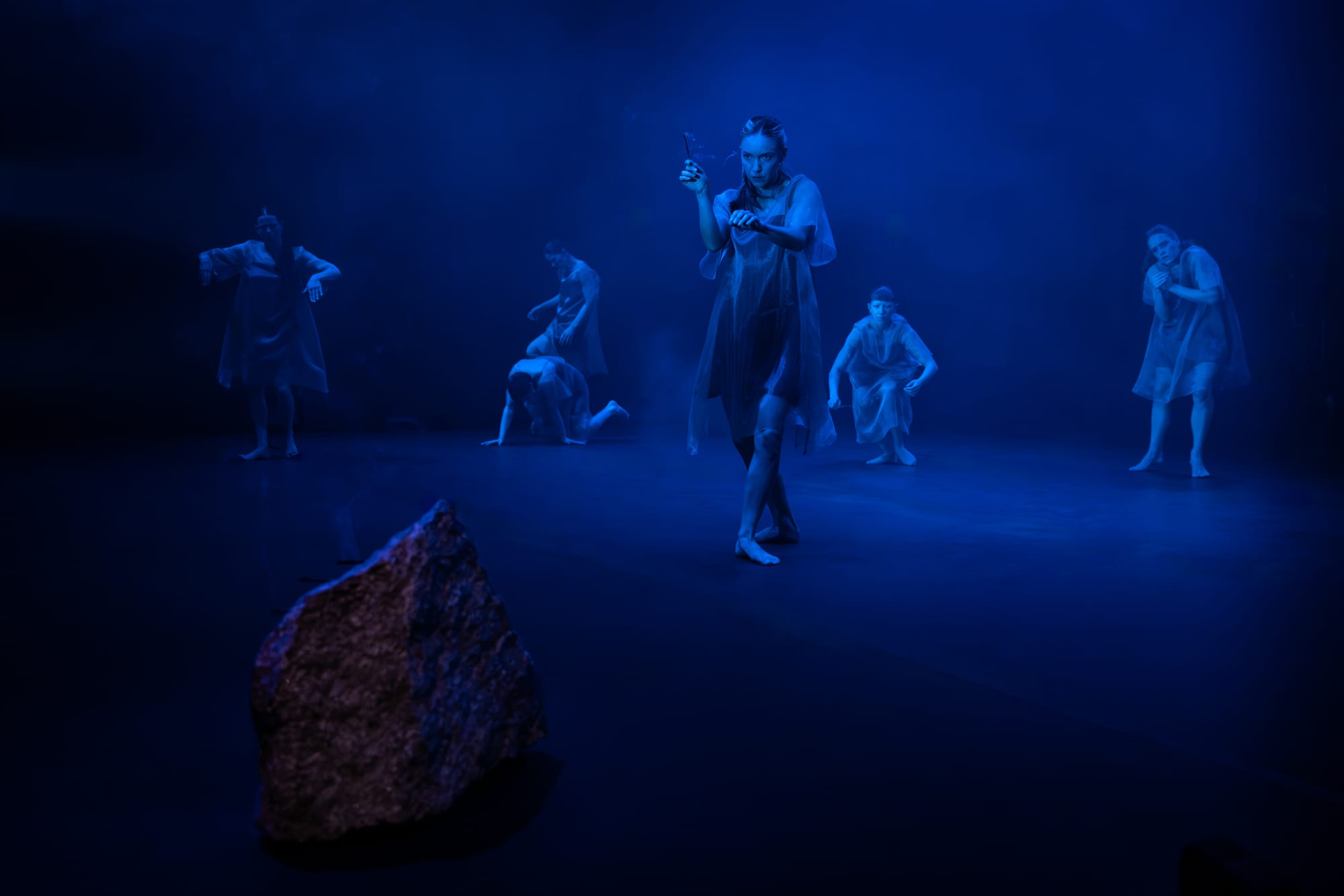 Trà Mi Dinh dances at centre stage under deep blue light, with other dancers visible through the haze behind her and a large rock in the foreground.