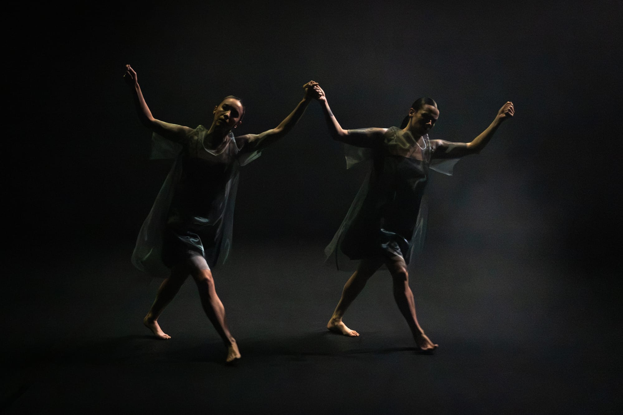 Trà Mi Dinh and Rachel Coulson dance side by side, arms raised and bodies leaning in mirrored movement against a dark, hazy stage.