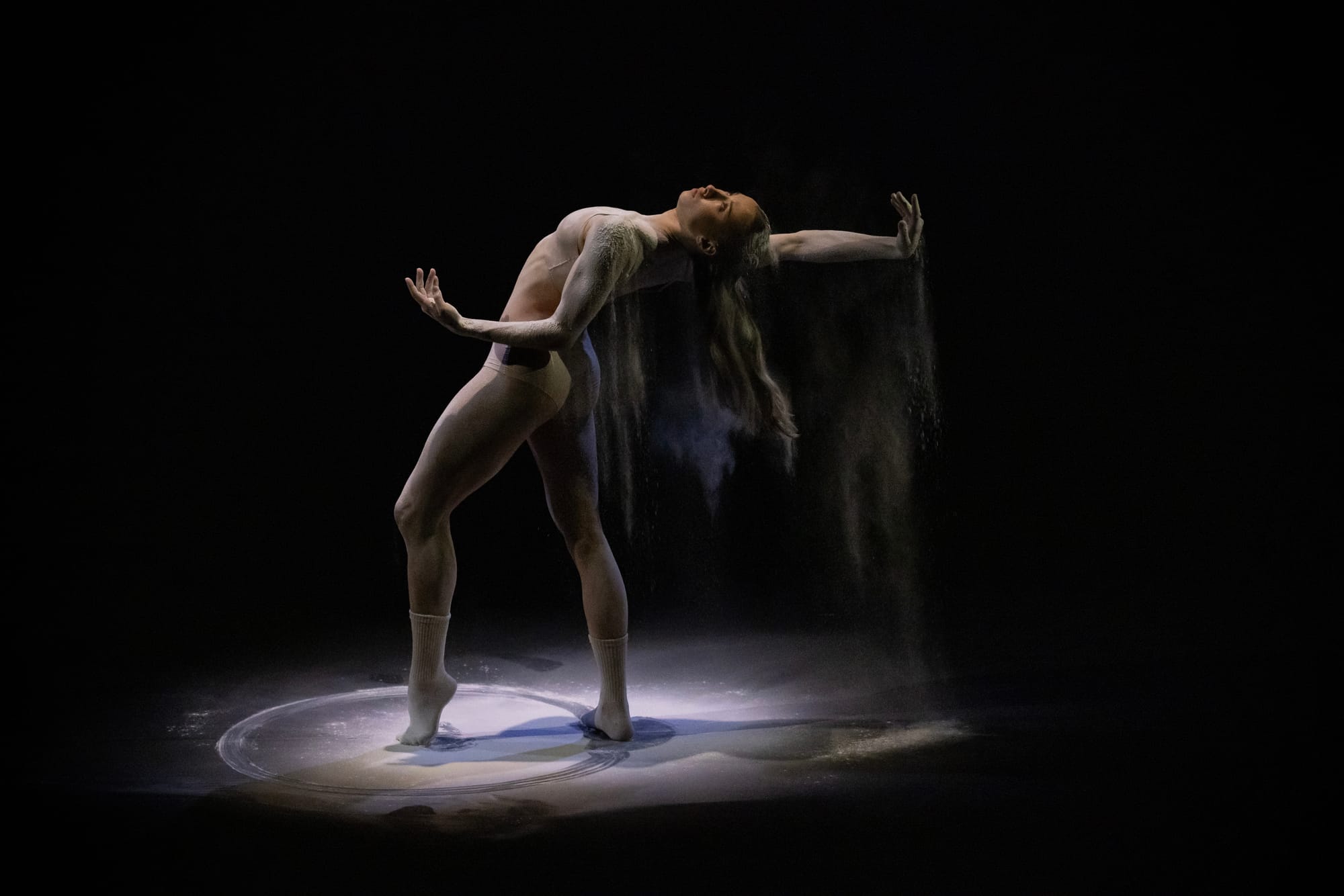 A dancer in a nude-toned costume arches backwards as flour falls from her hair and hands, gathering in sweeping circular traces on the black floor.