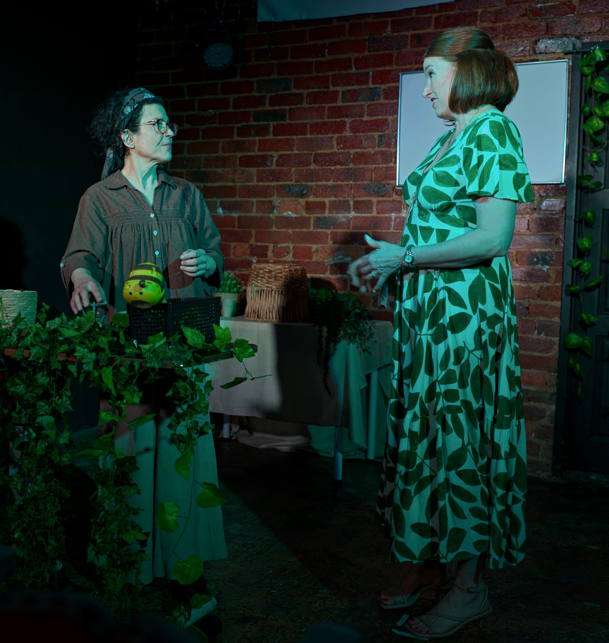 Nadia Andary stands behind a vine-covered counter facing Carissa McPherson, who wears a green-and-white patterned dress.