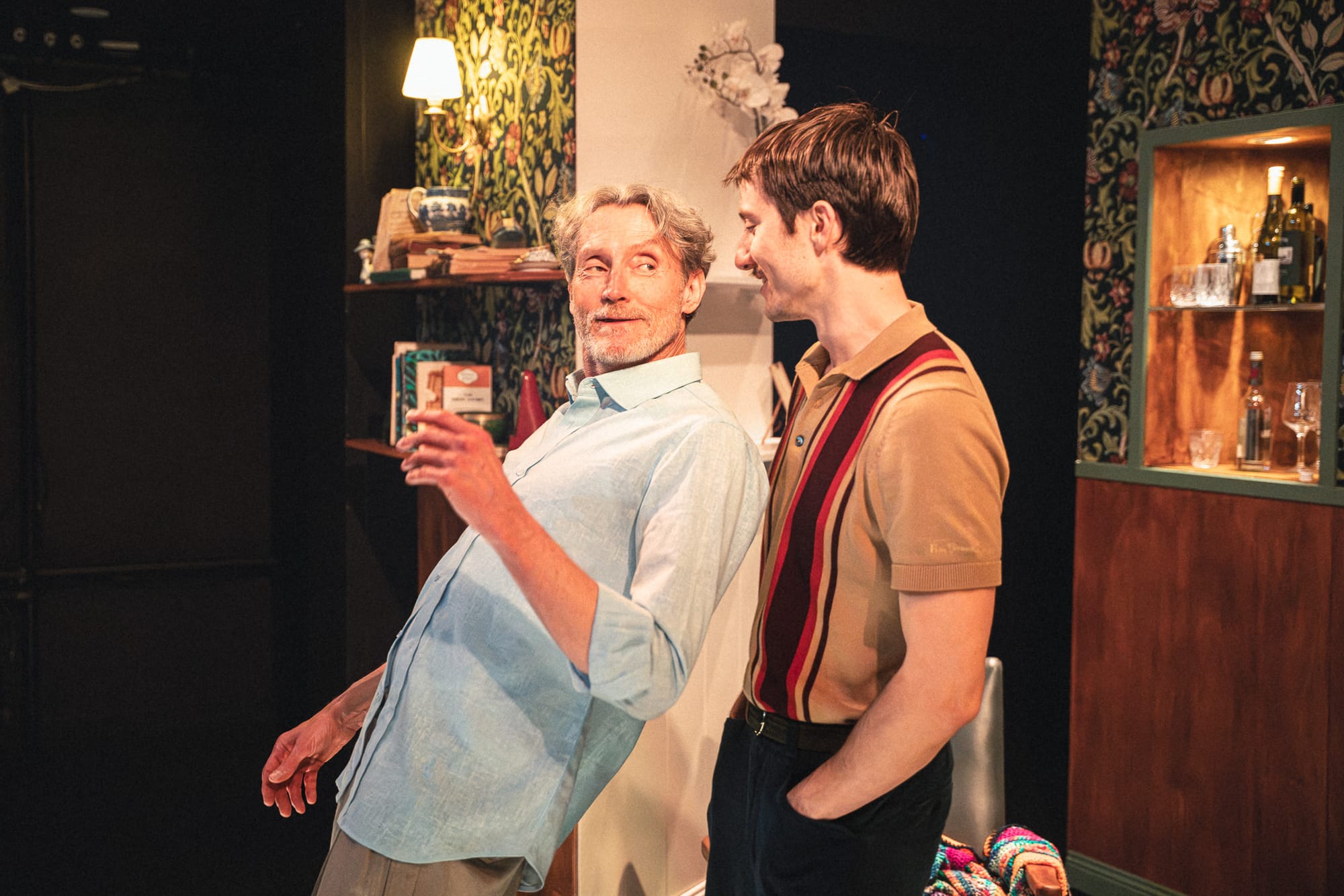 Dion Mills and Will Lonsdale stand very close together in the warm, patterned living room set, Mills leaning back playfully while holding a drink as Lonsdale watches him.