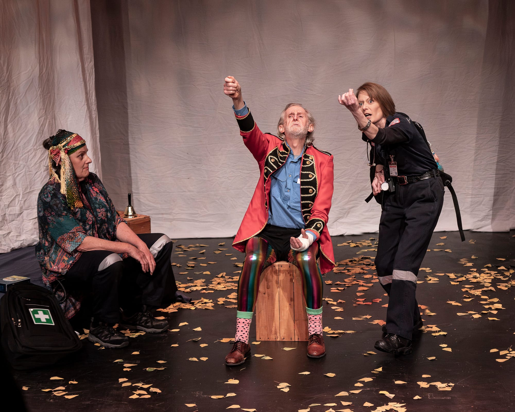 Anthea Davis sits at left, Don Bridges as Banjo sits centre in a red jacket and rainbow trousers, and Christine Husband stands at right in paramedic uniform on a stage scattered with paper leaves.
