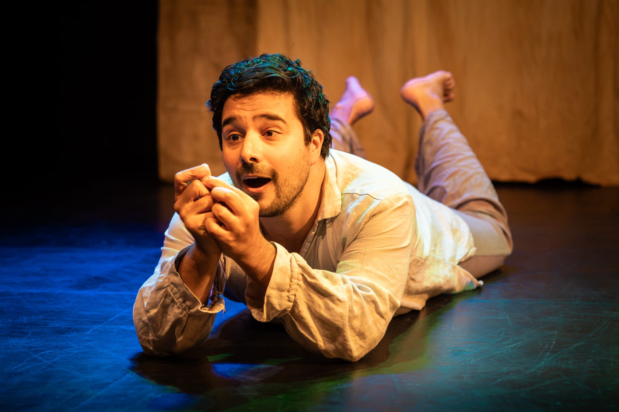 Fabio Motta in Holy Fool, lying on his stomach onstage, feet raised behind him as he gazes at something held closely in his hands.
