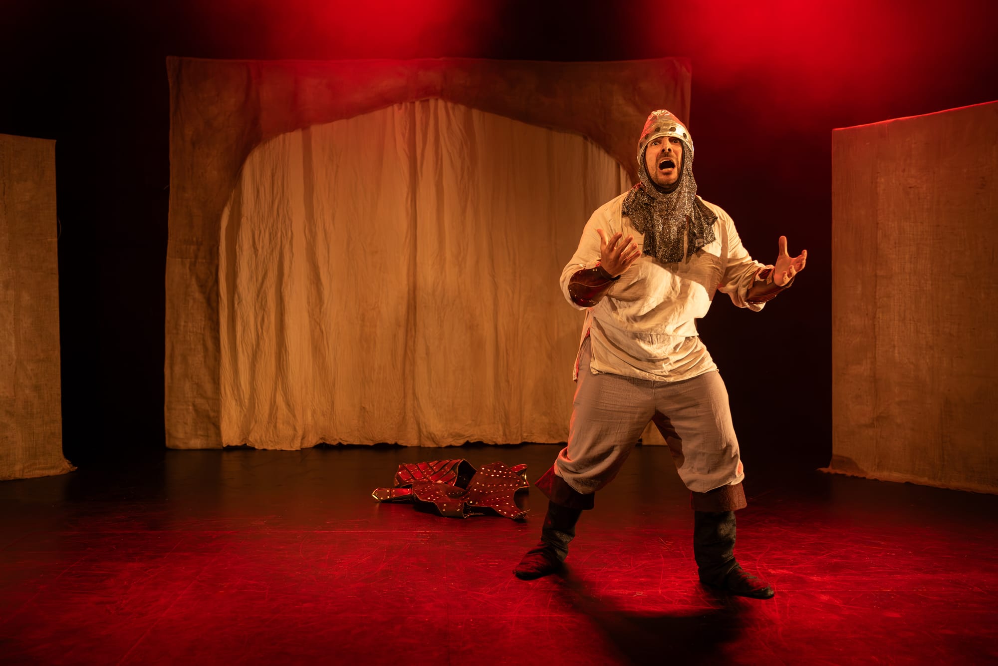 Fabio Motta in Holy Fool, dressed as a medieval soldier and standing centre stage beneath vivid red lighting.