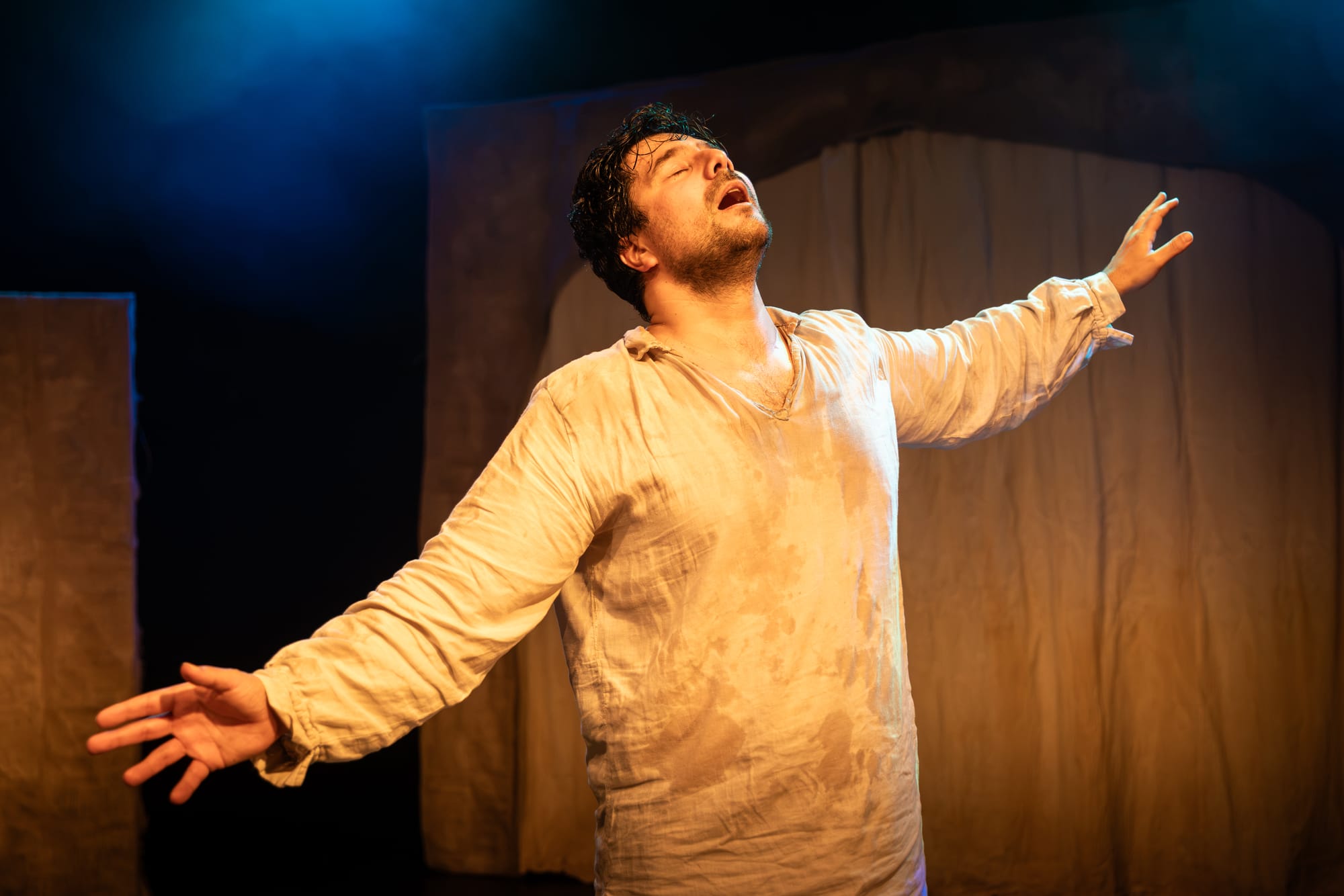 Fabio Motta in Holy Fool, barefoot with his arms spread wide and face lifted upwards in bliss, beneath blue and amber stage lighting.