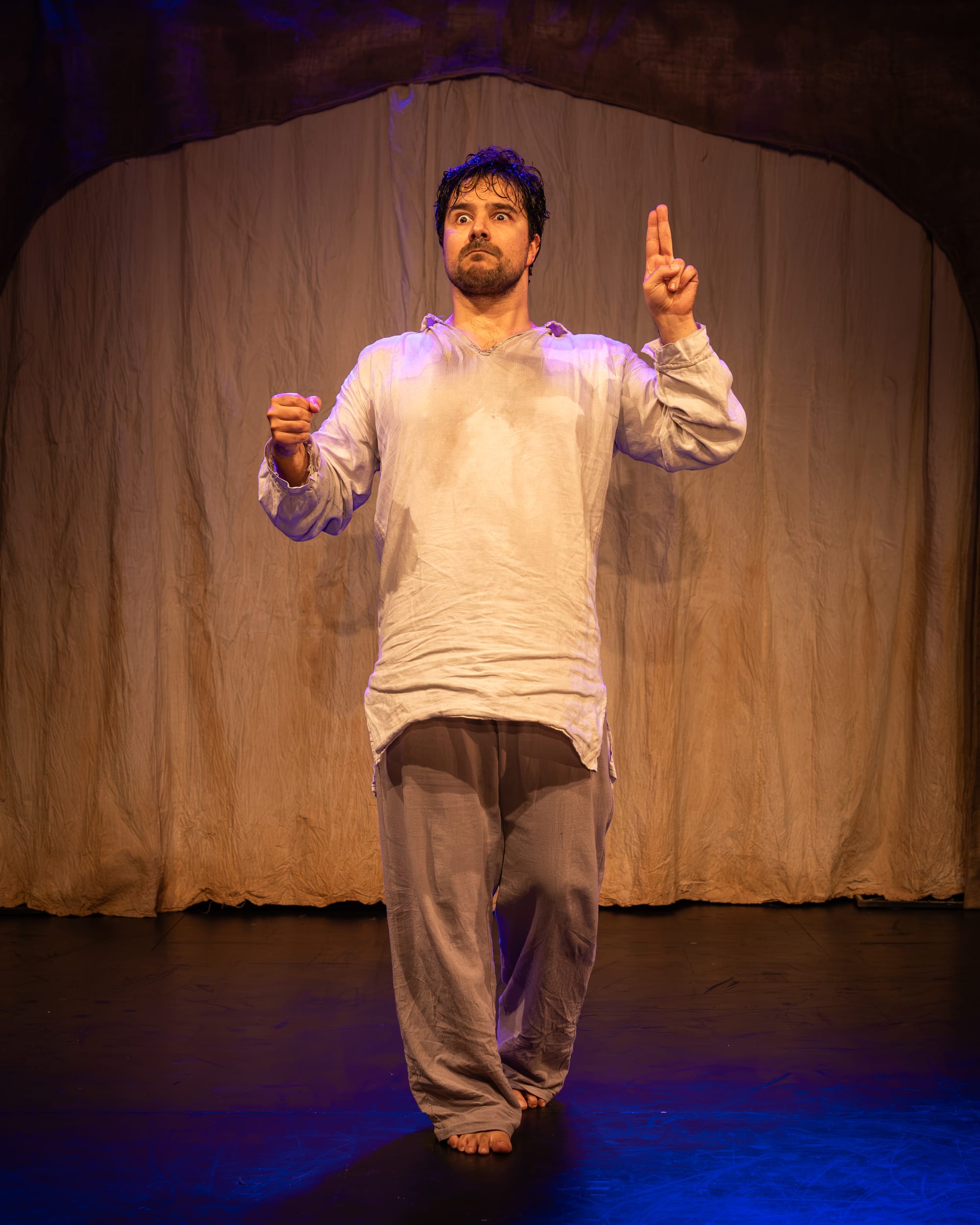 Fabio Motta in Holy Fool, standing barefoot before a pale fabric backdrop, poised to make the sign of the cross.