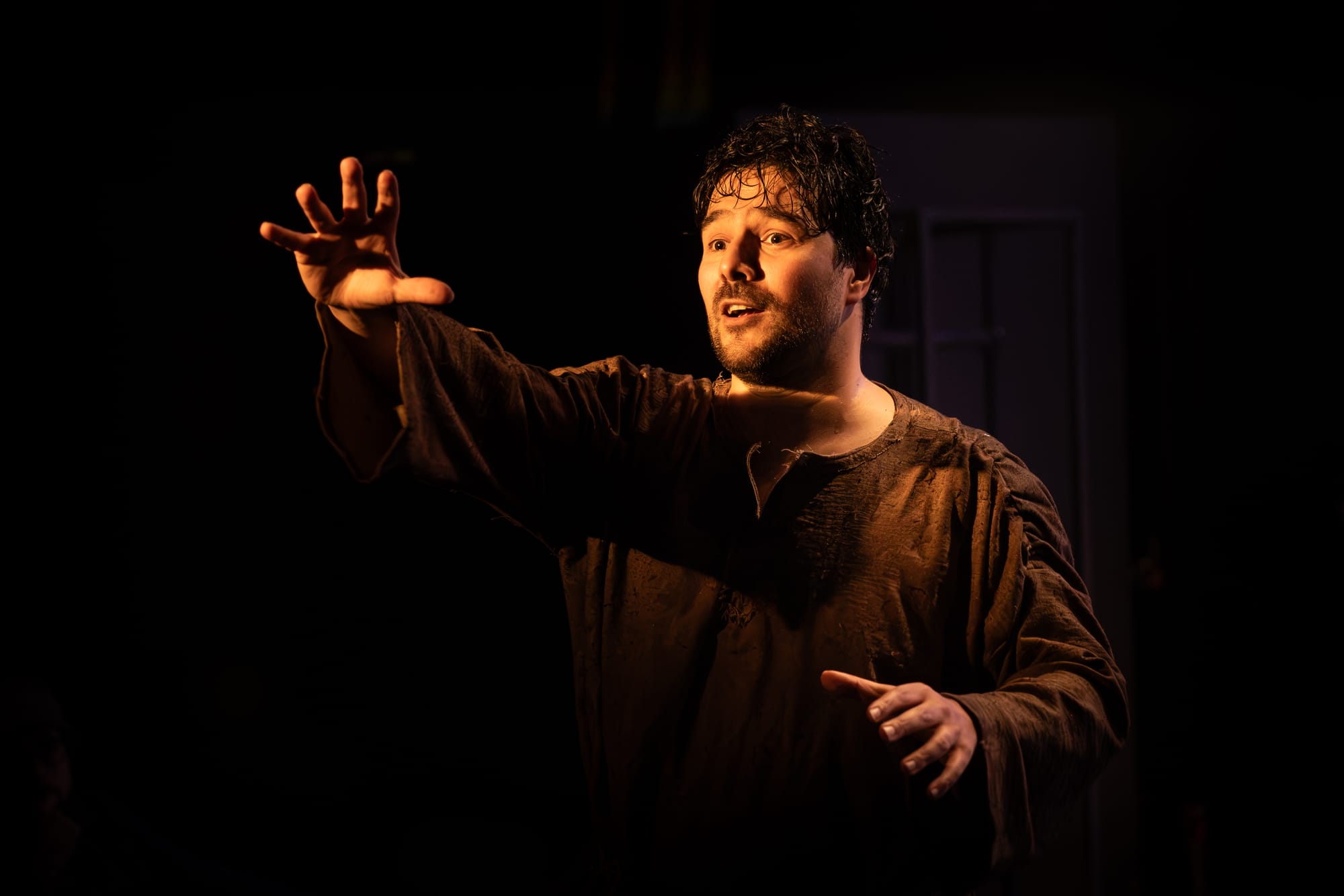 Fabio Motta in Holy Fool, wearing a dark brown robe and reaching one hand out into the darkness.
