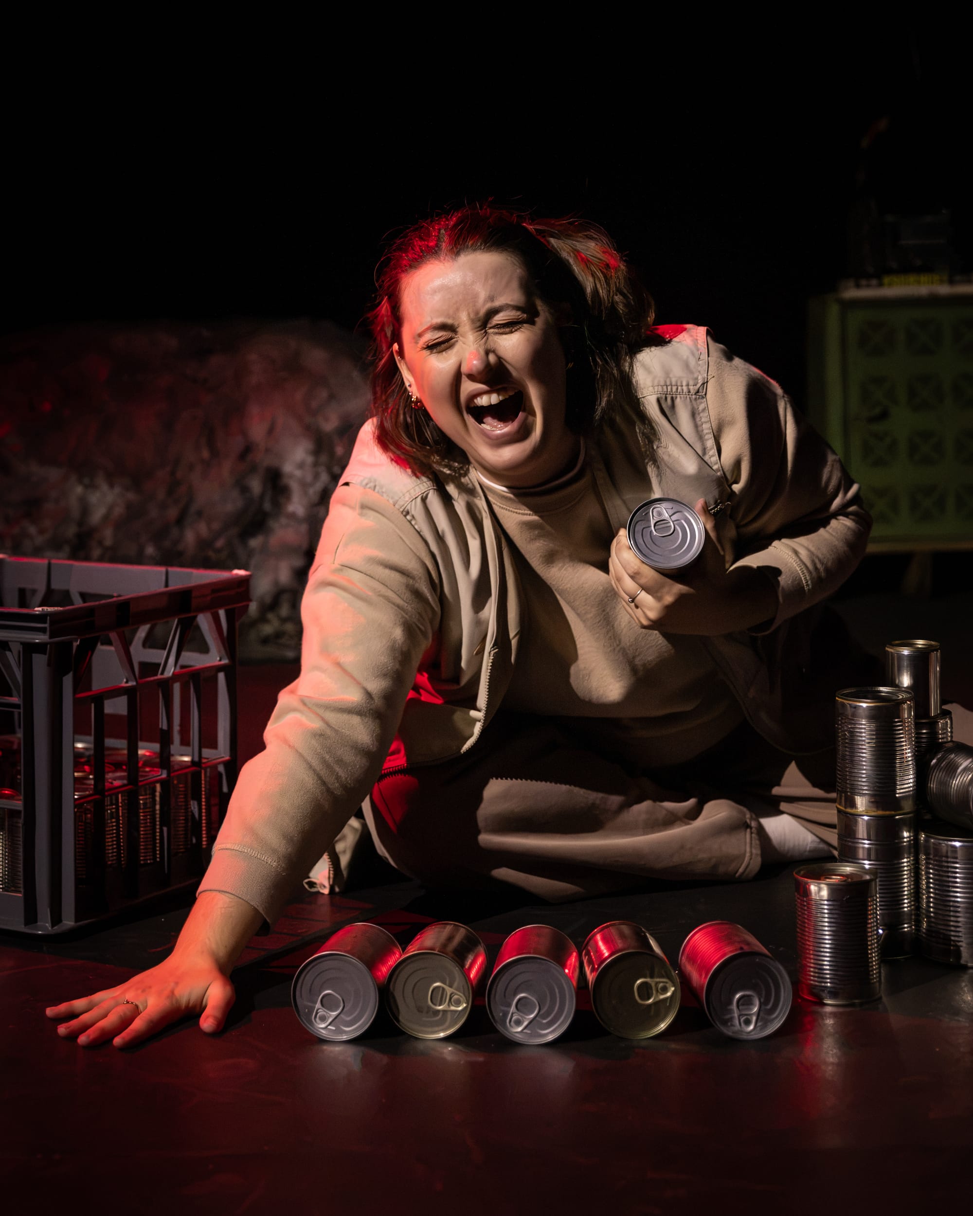 Jamila Main as Cam doubles over in pain among stacks of tins, one hand braced against the floor.