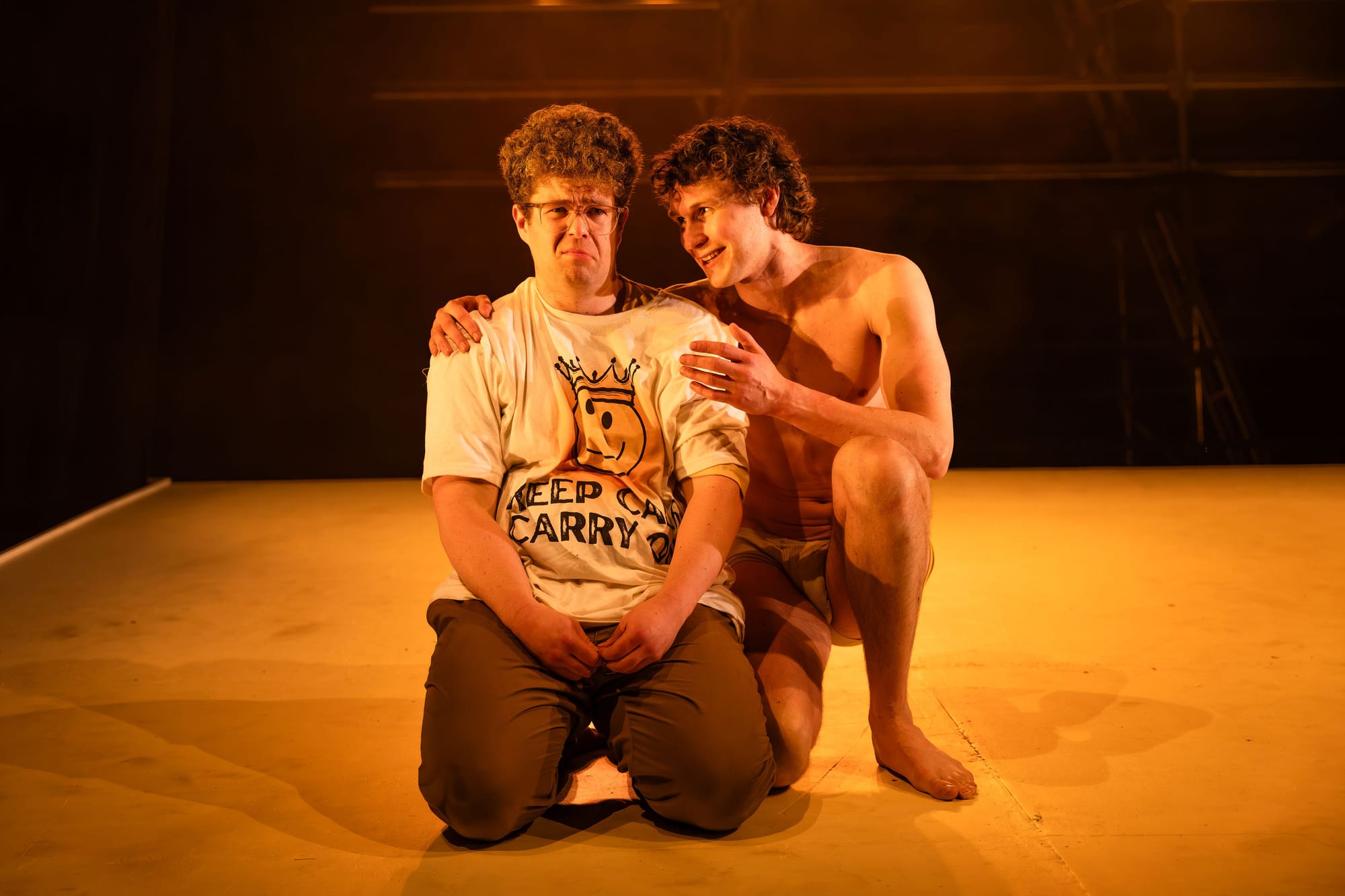 Oliver Cowen kneels at the front of the raked stage while Angus Leighton crouches closely behind him with one arm around his shoulder, speaking towards him under warm amber lighting.