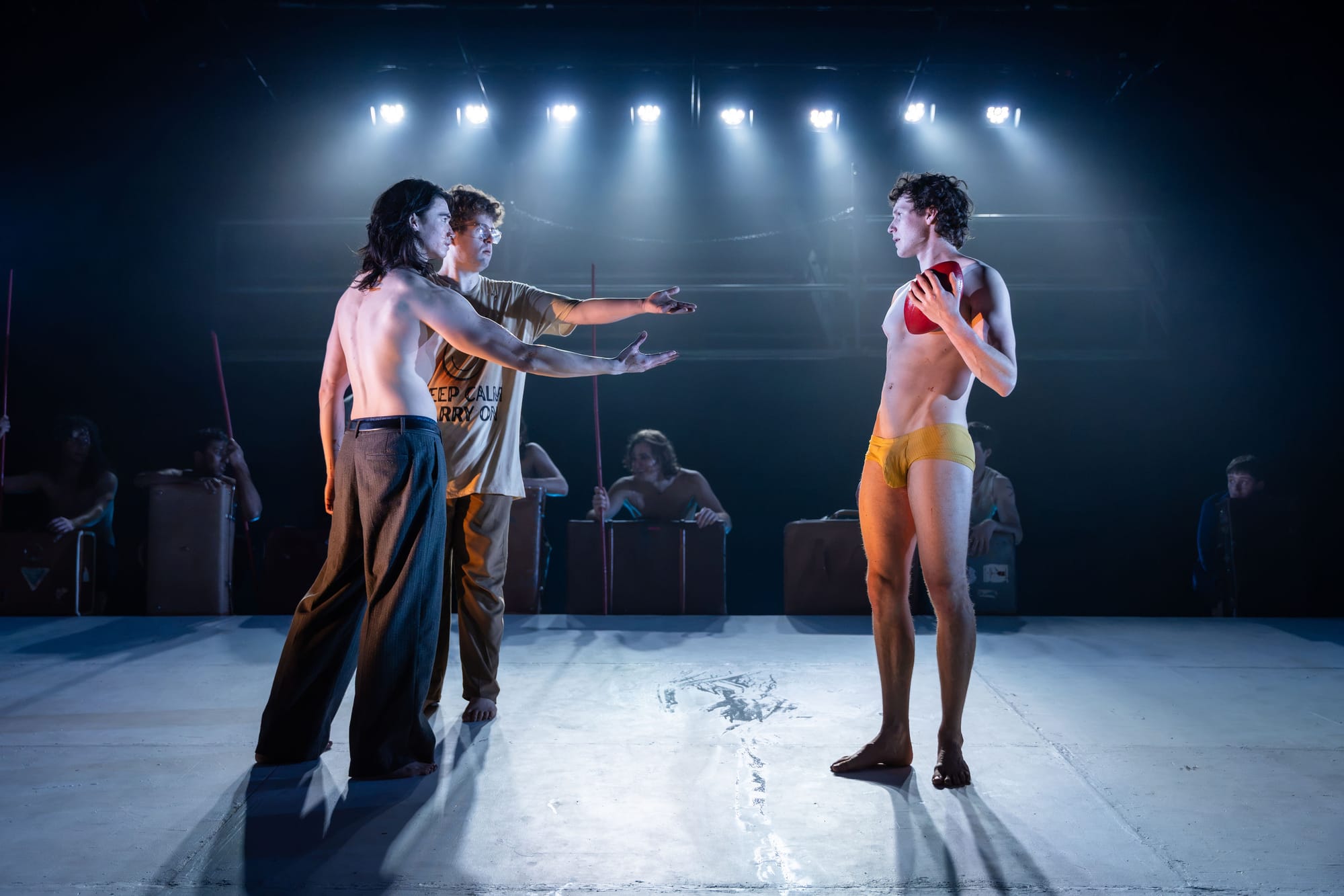Performers face one another on the brightly lit raked stage. Sean Yuen Halley and Oliver Cowen reach towards Angus Leighton, who stands apart in yellow briefs, holding an AFL football.
