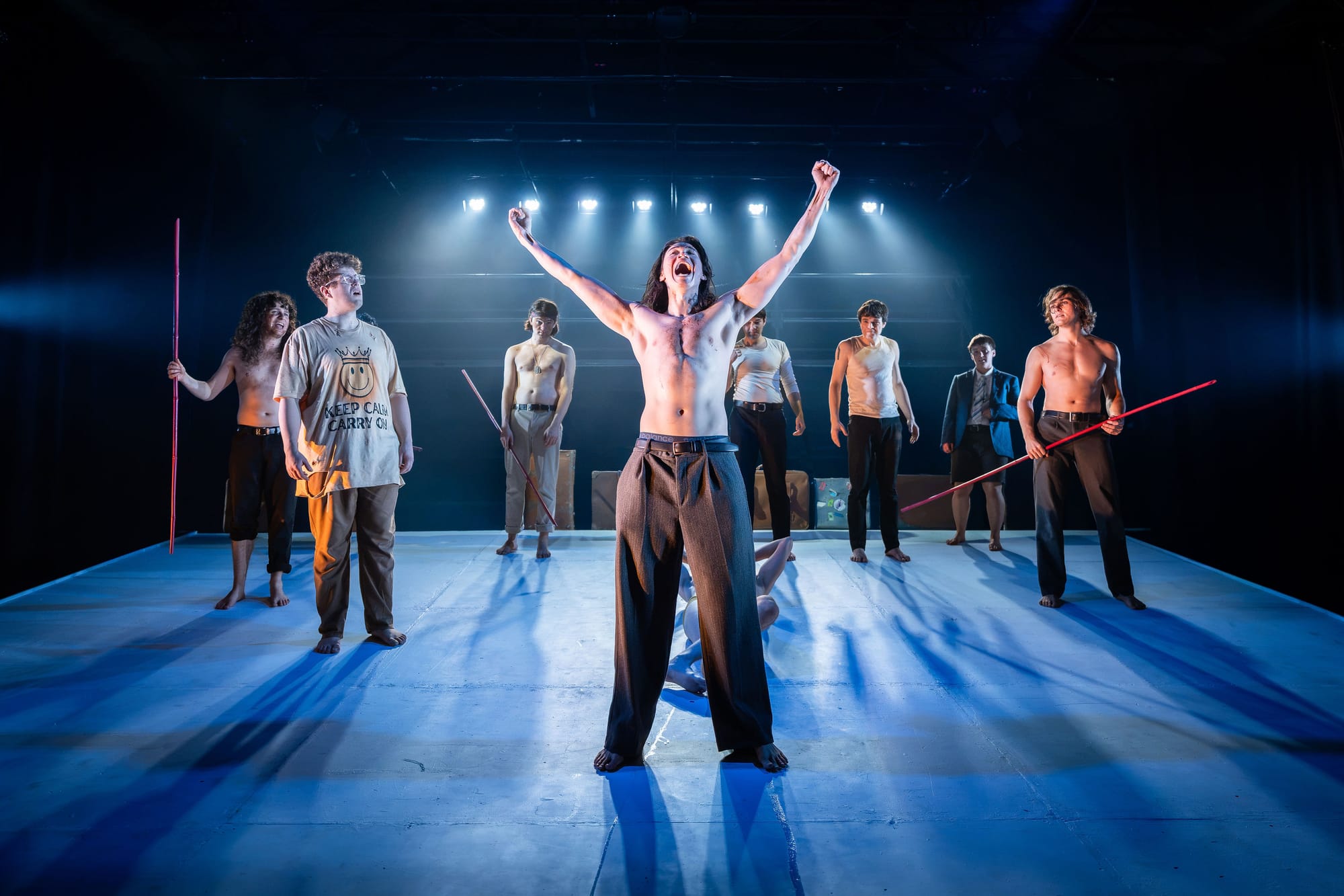 Sean Yuen Halley stands centre stage, bare-chested with both arms raised, surrounded by the Lord of the Flies ensemble across the rake. Several performers hold long red poles beneath stark blue-white lighting.