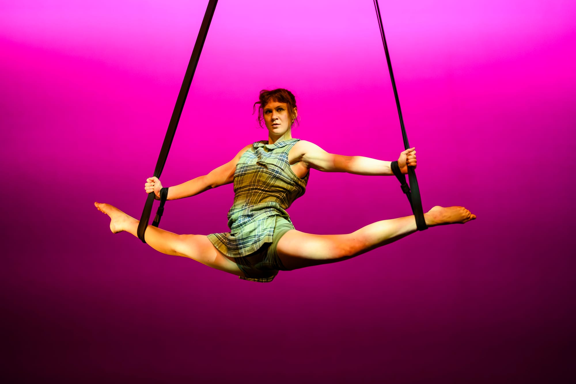 Lara Elliot performs an aerial split on straps high above the floor against vivid pink lighting.