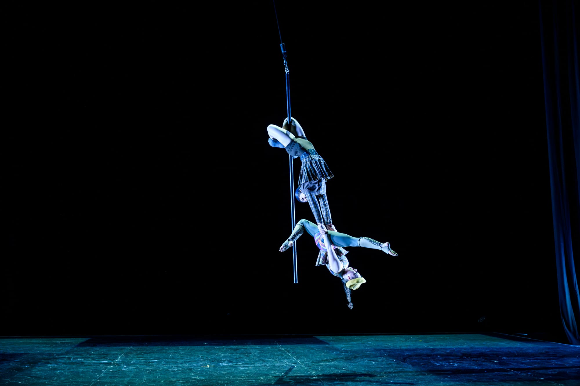 Capri Palmer and Shayla Ashton perform high above the circus floor on the swinging pole under blue lighting.