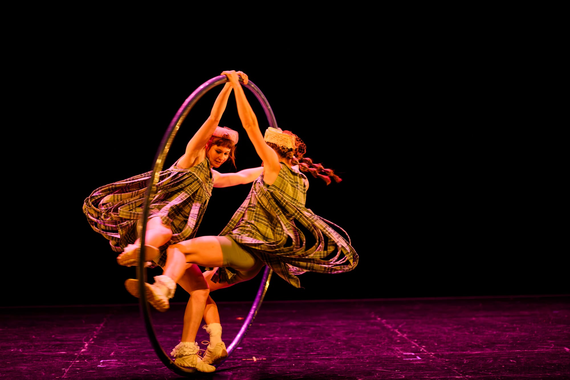 Lara Elliot and Nina Smith perform together inside a Cyr wheel under pink stage lighting.