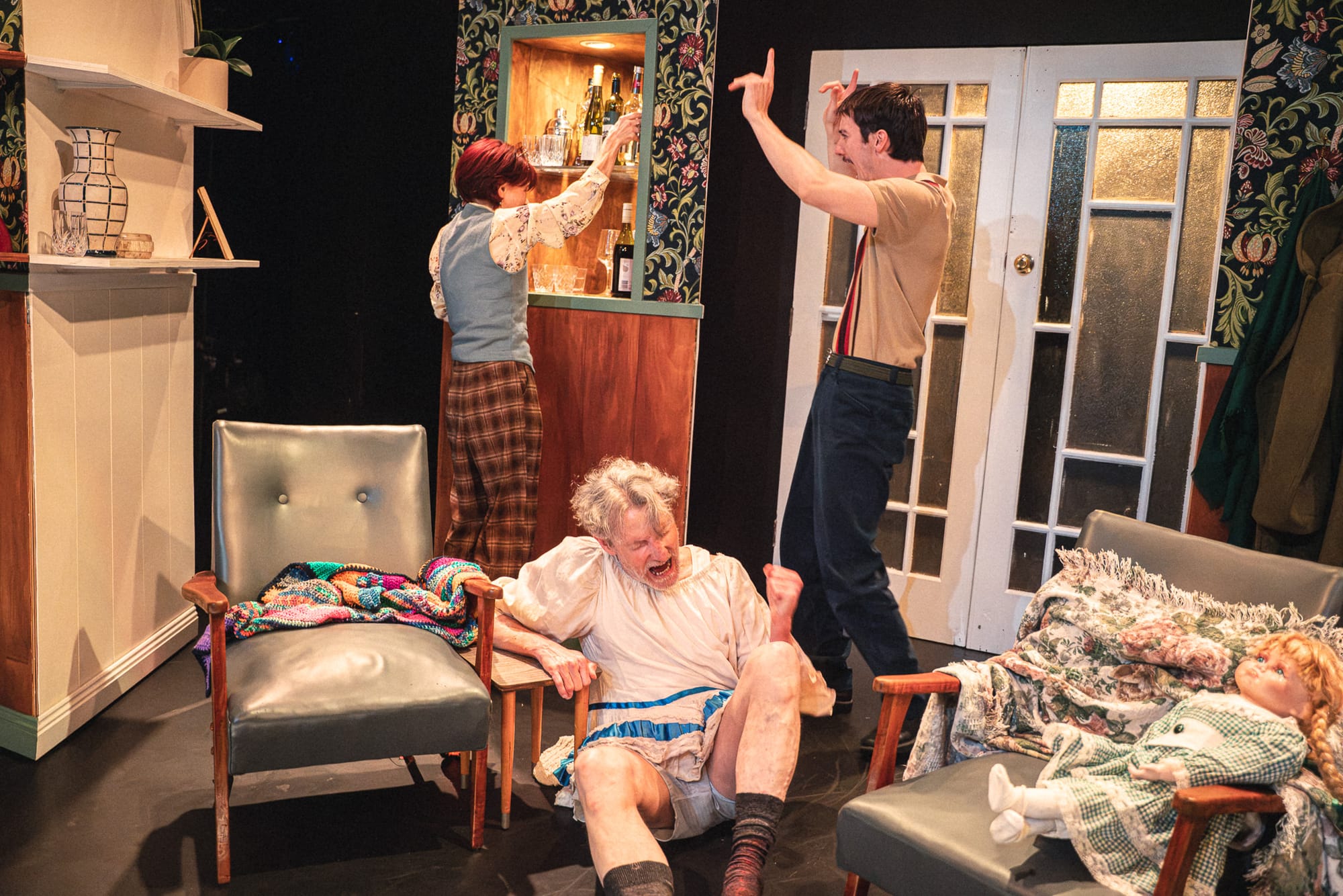 Dion Mills sits dramatically on the floor in a pale dress while Will Lonsdale gestures behind him and Olga Makeeva stands near the drinks cabinet. A large doll lies across an armchair beside them.