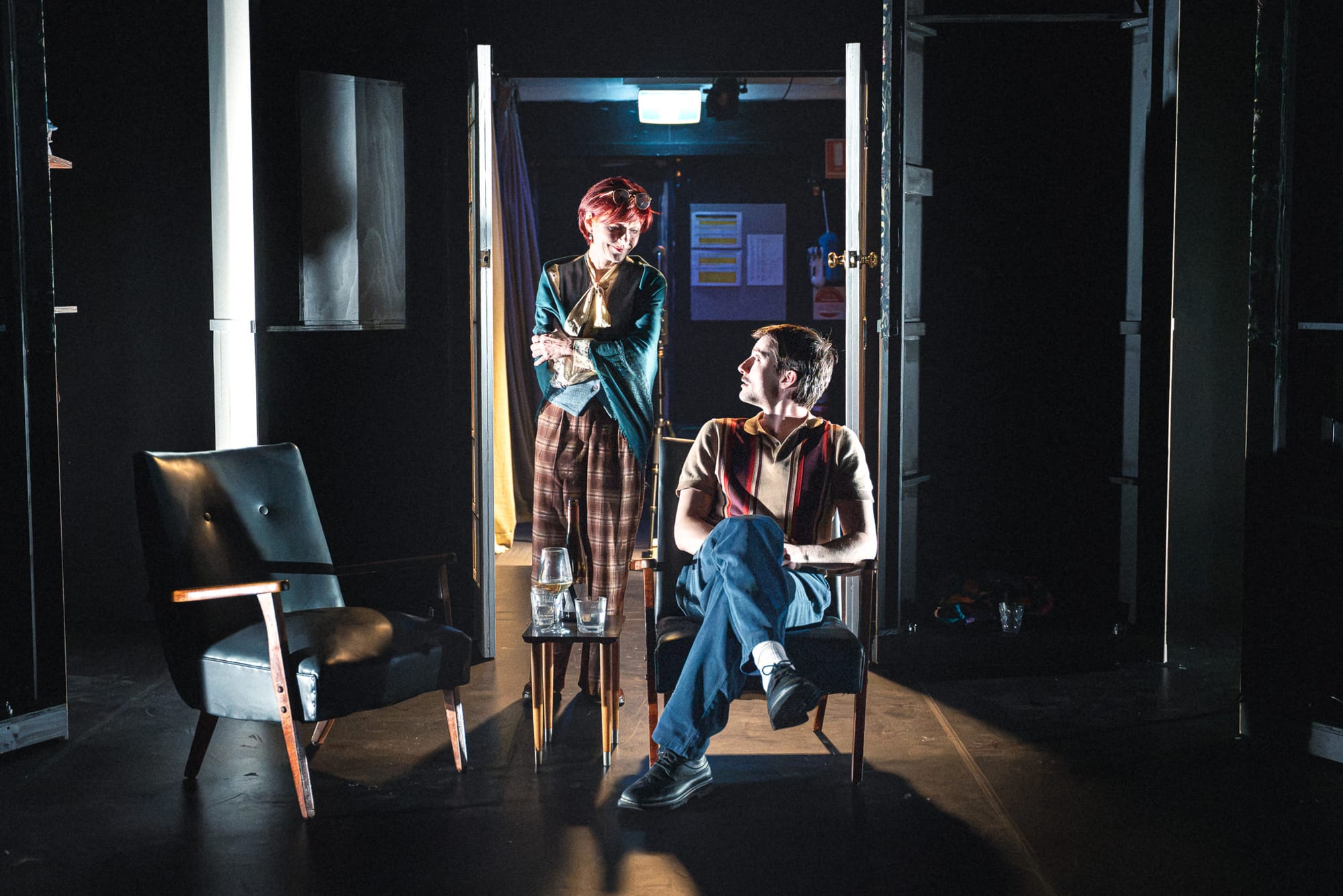 Olga Makeeva stands over a seated Will Lonsdale in a dark, exposed version of the set, with the backs of scenic structures and an open backstage doorway visible around them.