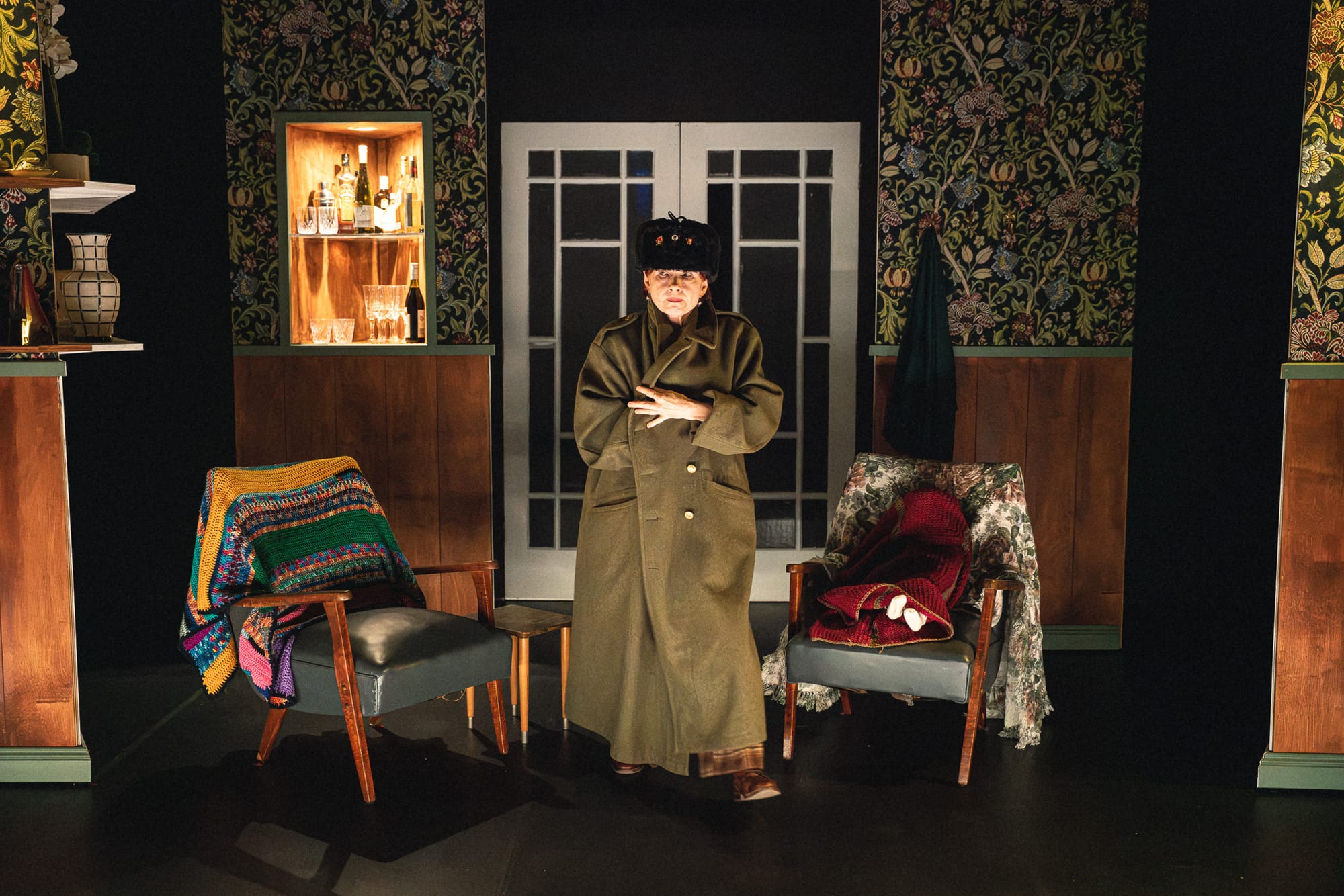 Olga Makeeva stands centre stage in an oversized dark military-style coat and large black fur hat, surrounded by patterned wallpaper, mismatched chairs and colourful crochet blankets.