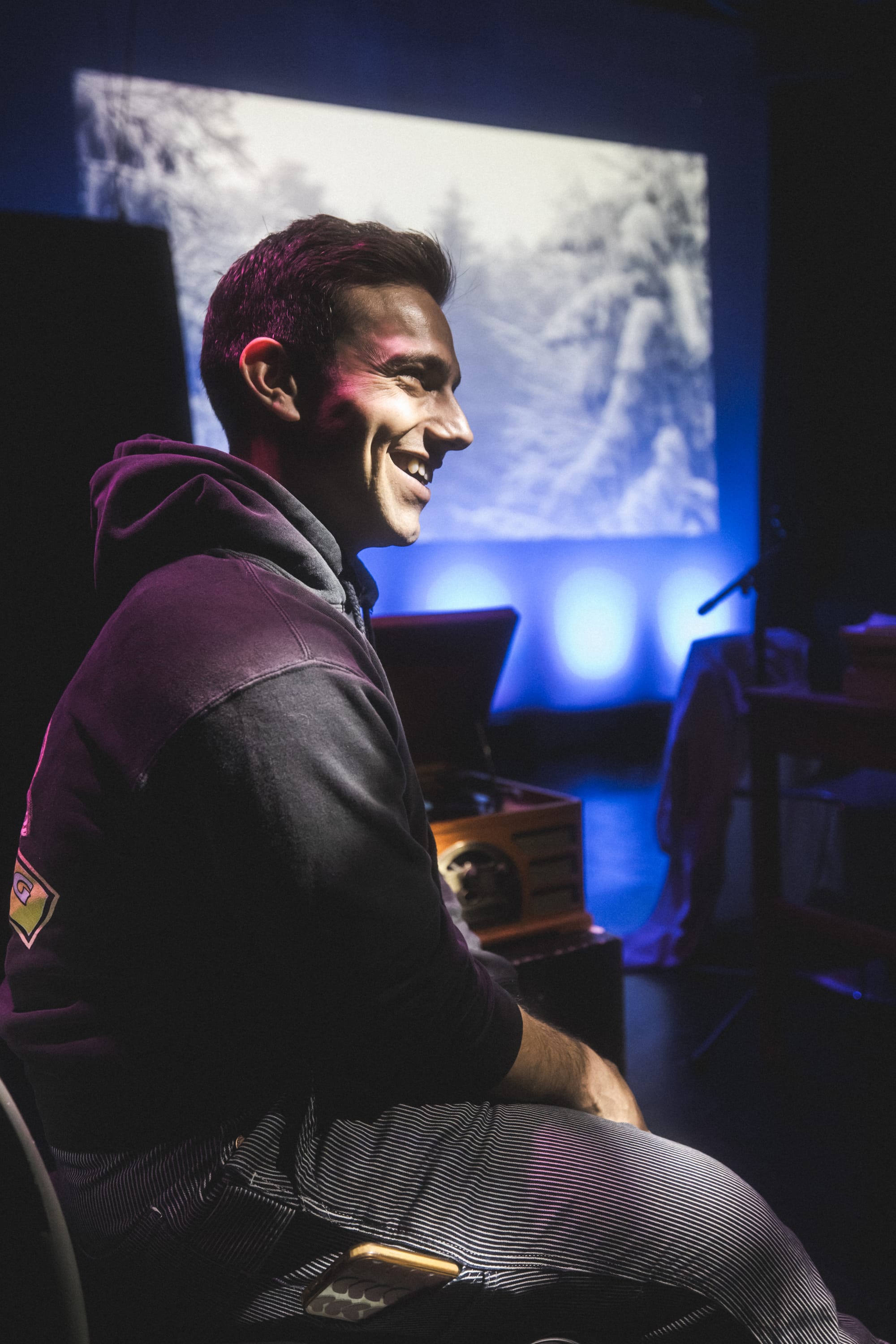 Gregory Nussen smiles in profile, with a large projected image and blue stage lighting behind them.