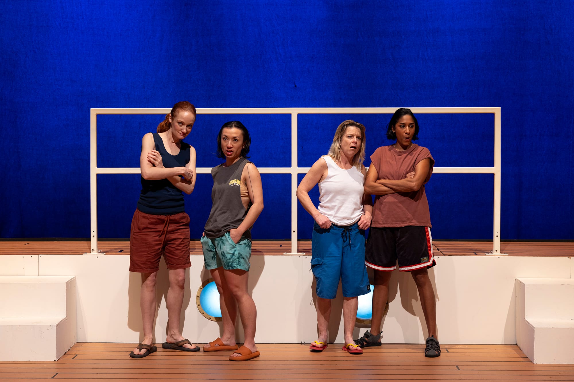 Four performers stand together on the white deck of a cruise ship against a vivid blue backdrop, dressed in casual men’s clothing.