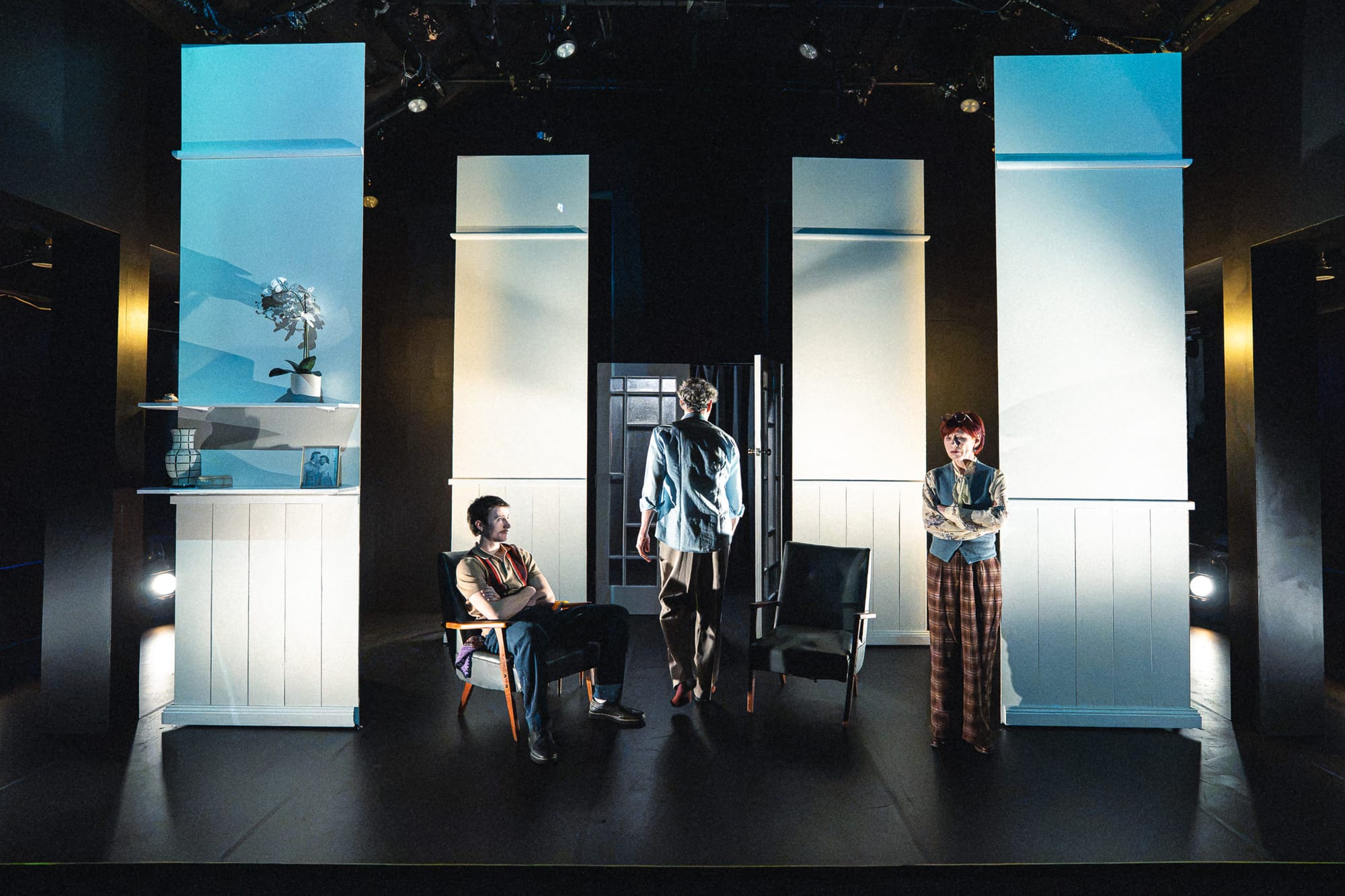 The three performers occupy a stark, mostly white version of the room, framed by tall pale scenic panels. Will Lonsdale sits at left, Dion Mills stands near the centre doorway and Olga Makeeva stands at right.