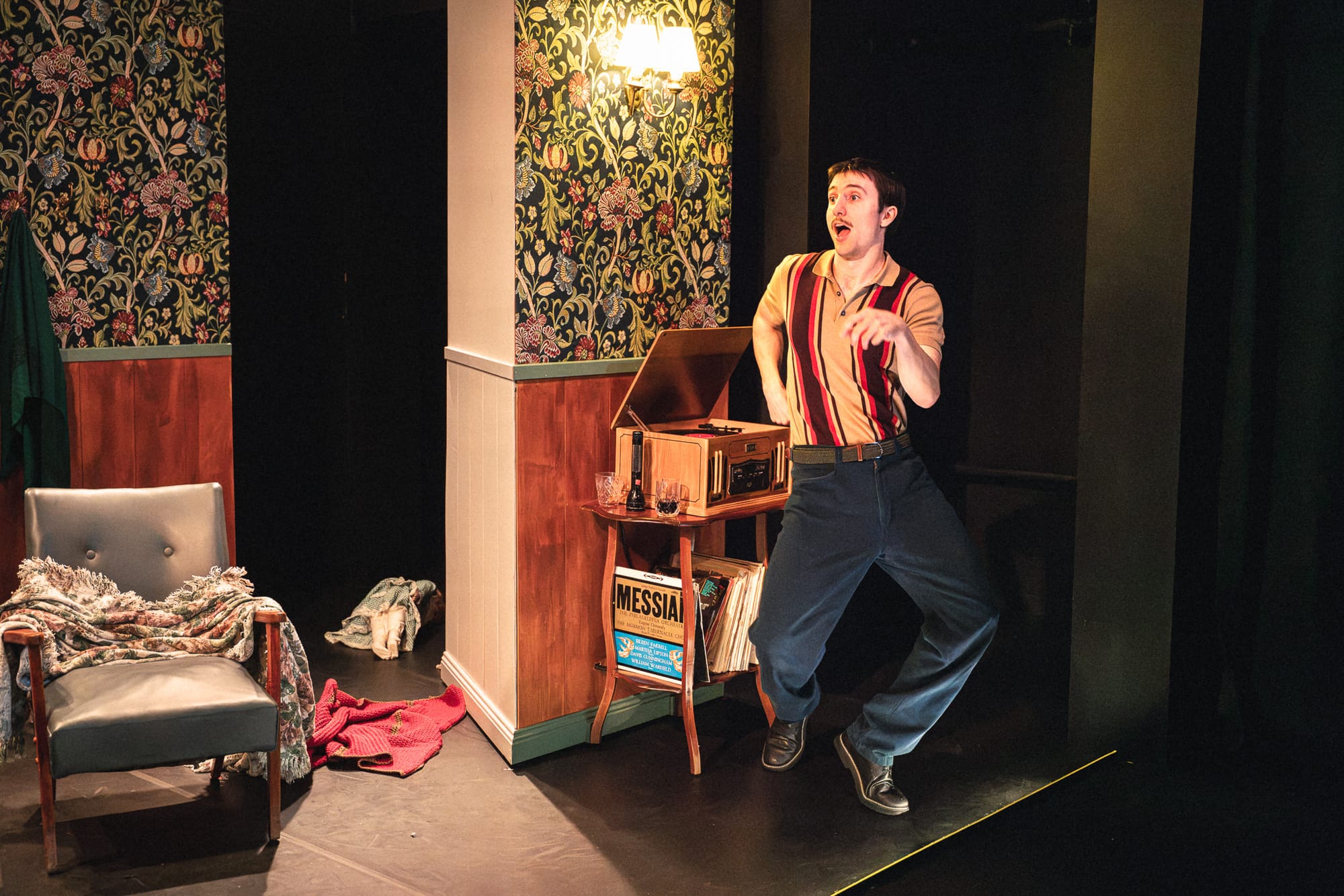 Will Lonsdale gestures animatedly beside a wooden record player in the richly patterned 2007 living room, with records stacked on the shelf below.