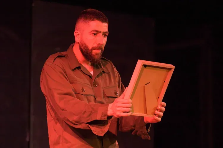 Mohammed El Kesm as Aviv, dressed in military uniform, looks at a framed photograph of Anne Frank under red stage lighting in The Return.